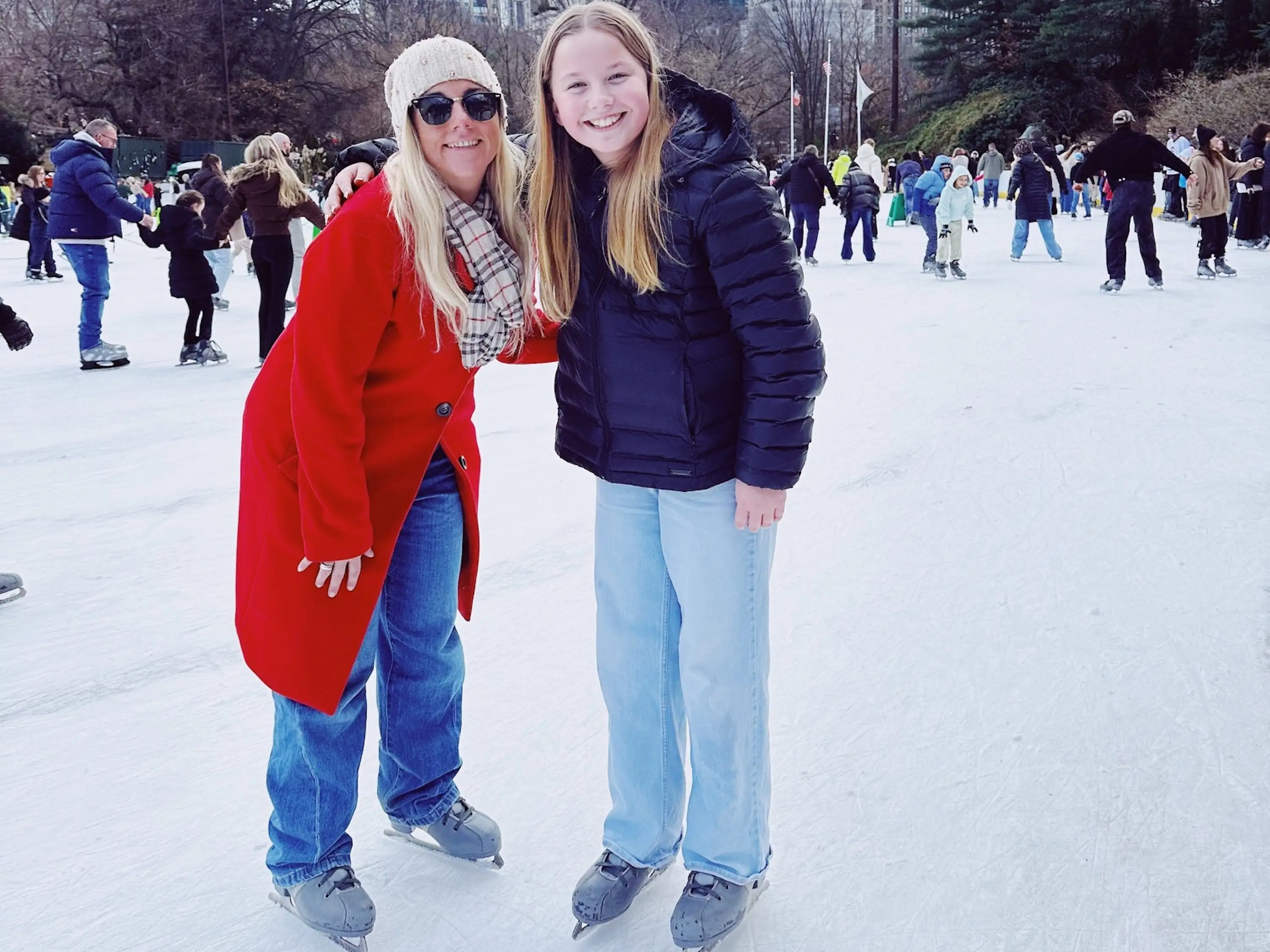 Two people wearing ice skates pose on a crowded outdoor rink beneath Manhattan skyscrapers.