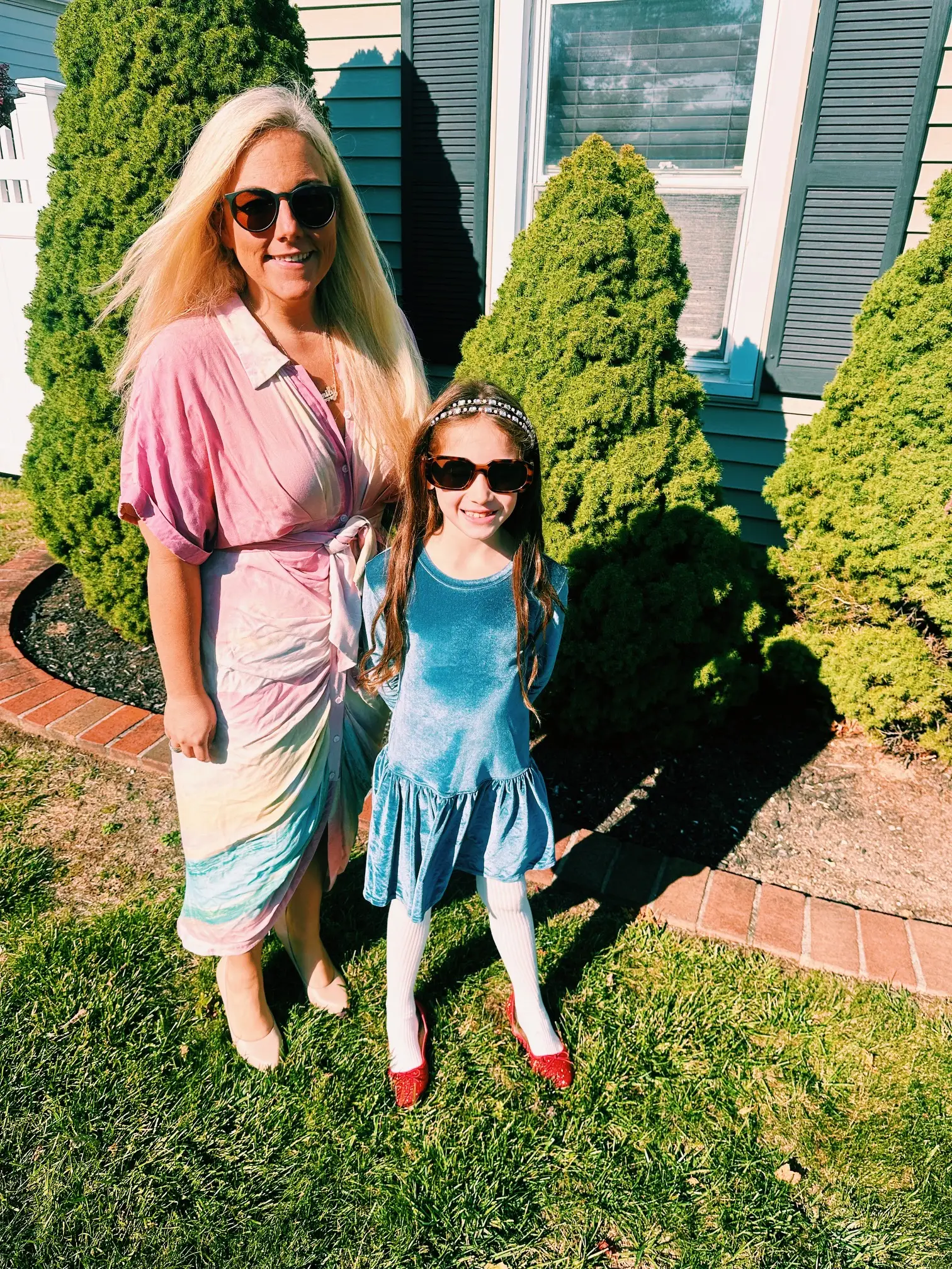 Two people wearing sunglasses stand on a sunny lawn beside trimmed shrubs and a house.