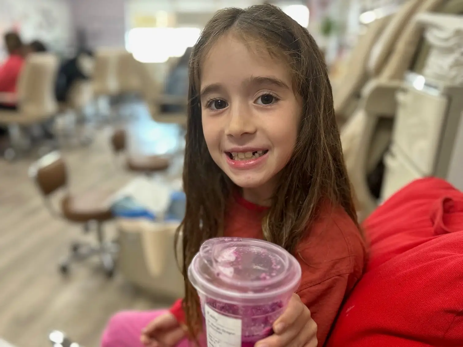 Person seated in a salon chair holds a clear cup of purple drink beside pedicure stations.