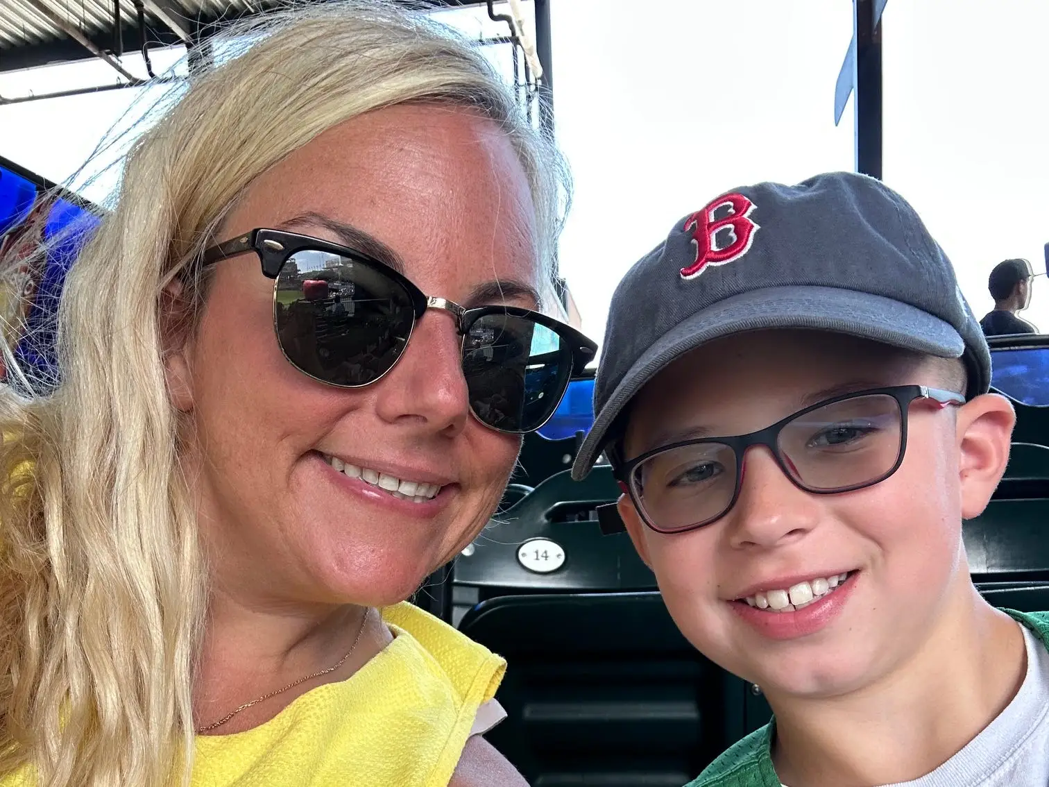 Two smiling spectators sit in stadium seats wearing sunglasses, glasses, and Boston Red Sox apparel.