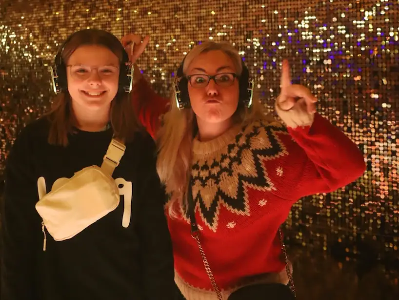 Two people wearing headphones pose in a gold sequined room with metallic fringe decorations overhead.