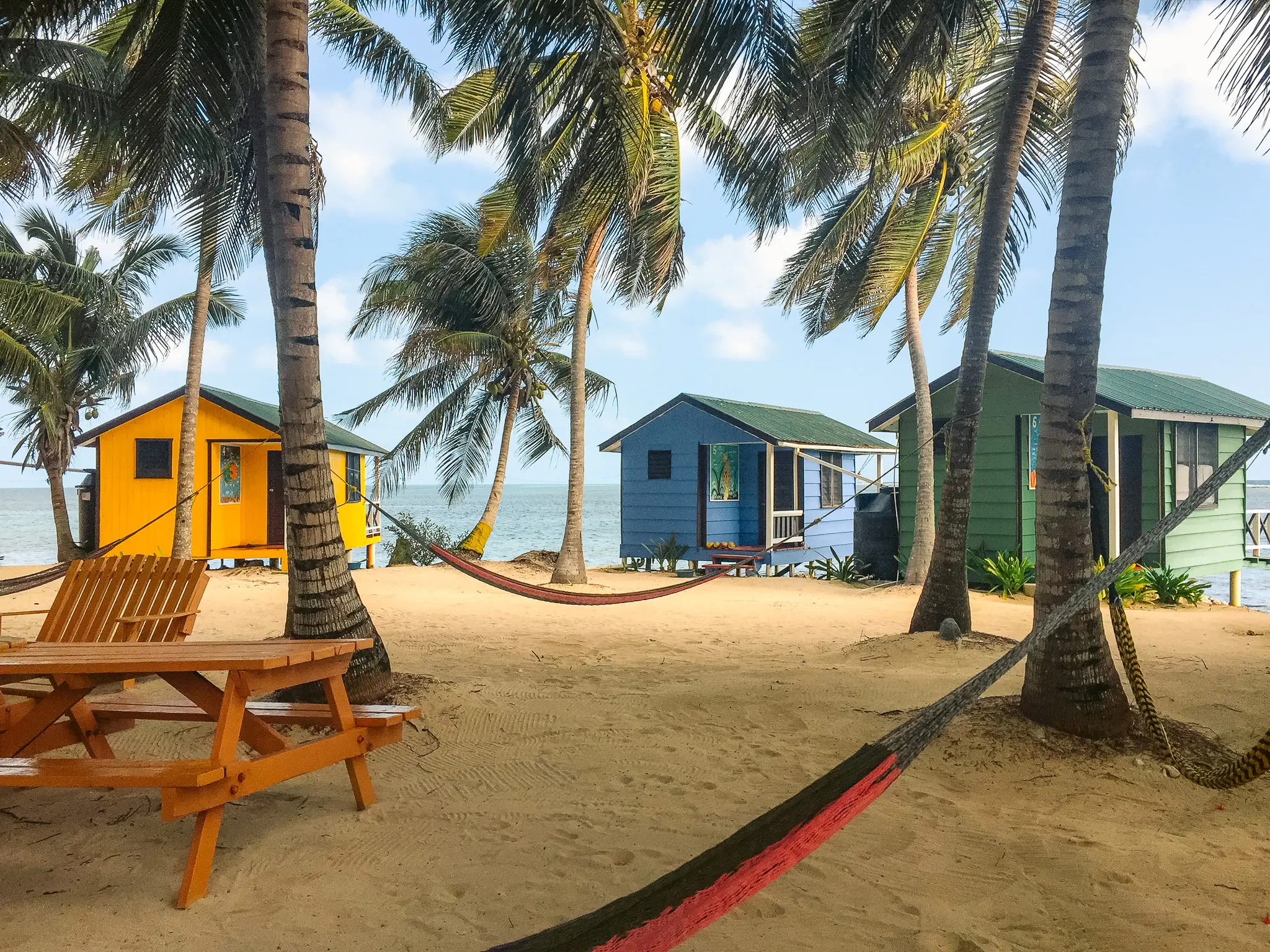 Three colorful shacks under palm trees on a beach.