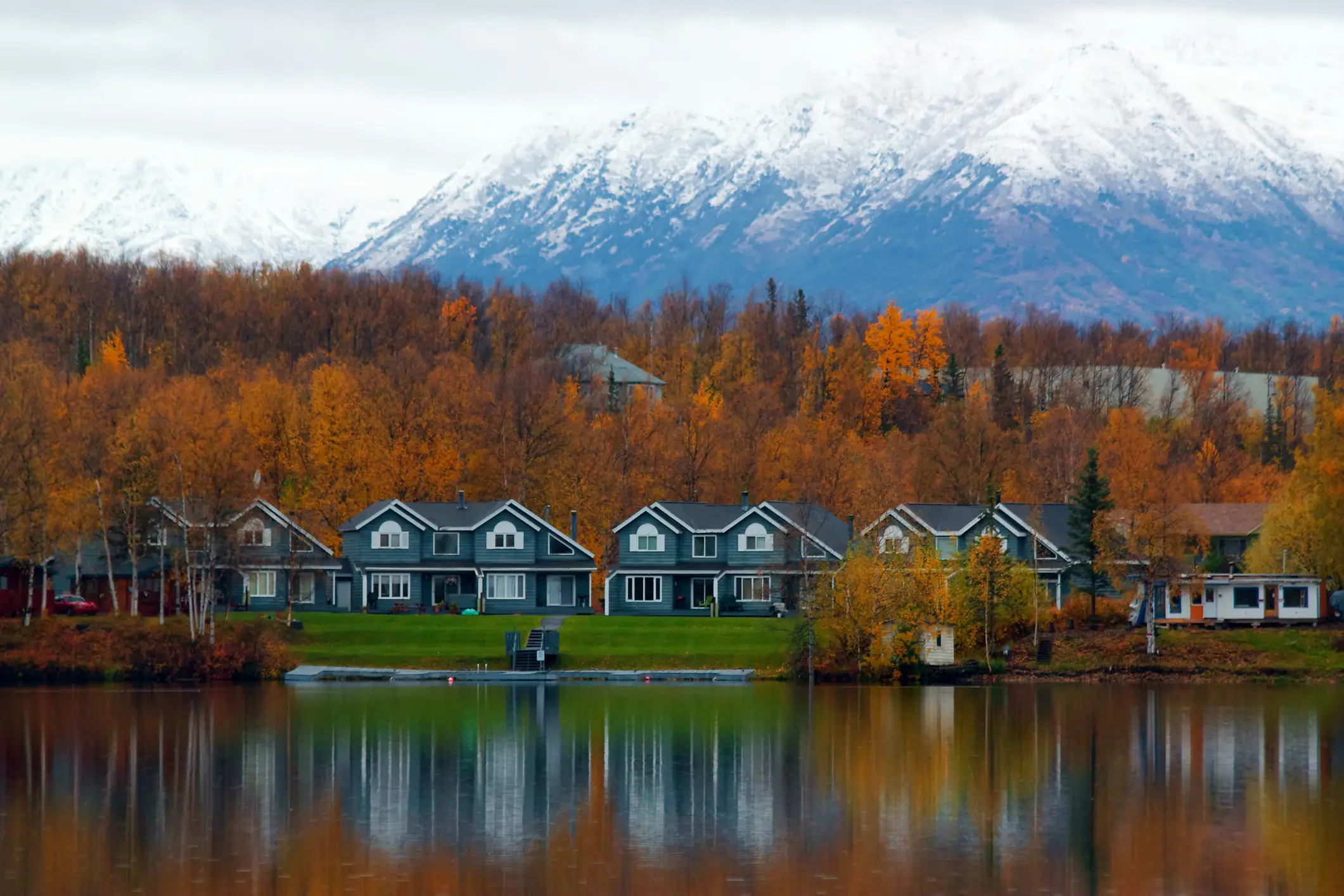 Lakeside homes reflect in calm water below autumn trees and snow-covered mountains.