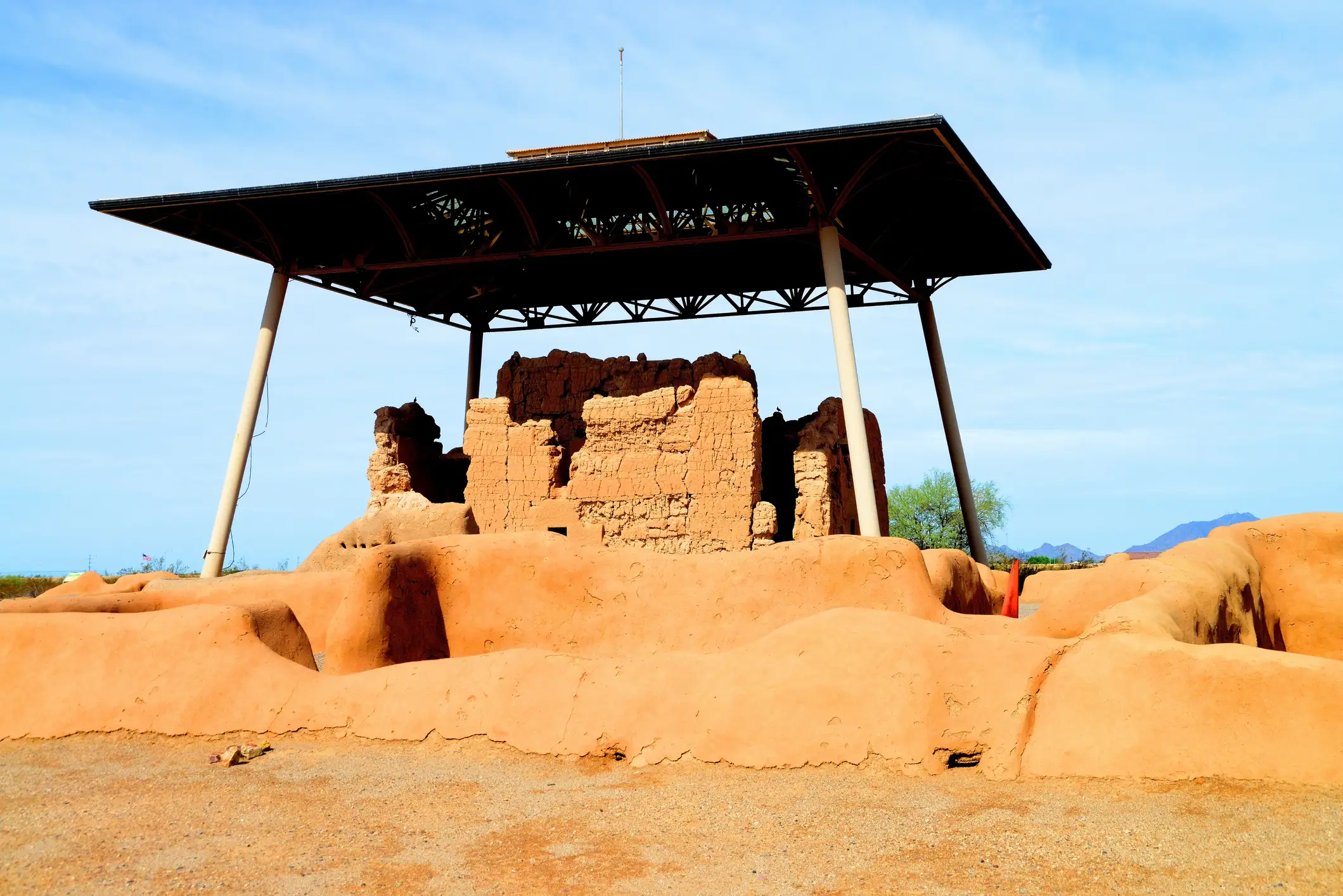 Adobe ruins stand beneath a large protective metal canopy in a sunny desert landscape.