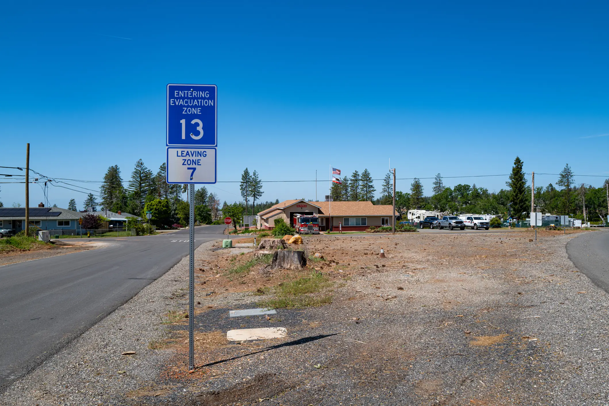 Evacuation zone signs stand beside a rural road near a fire station and cleared gravel lot.