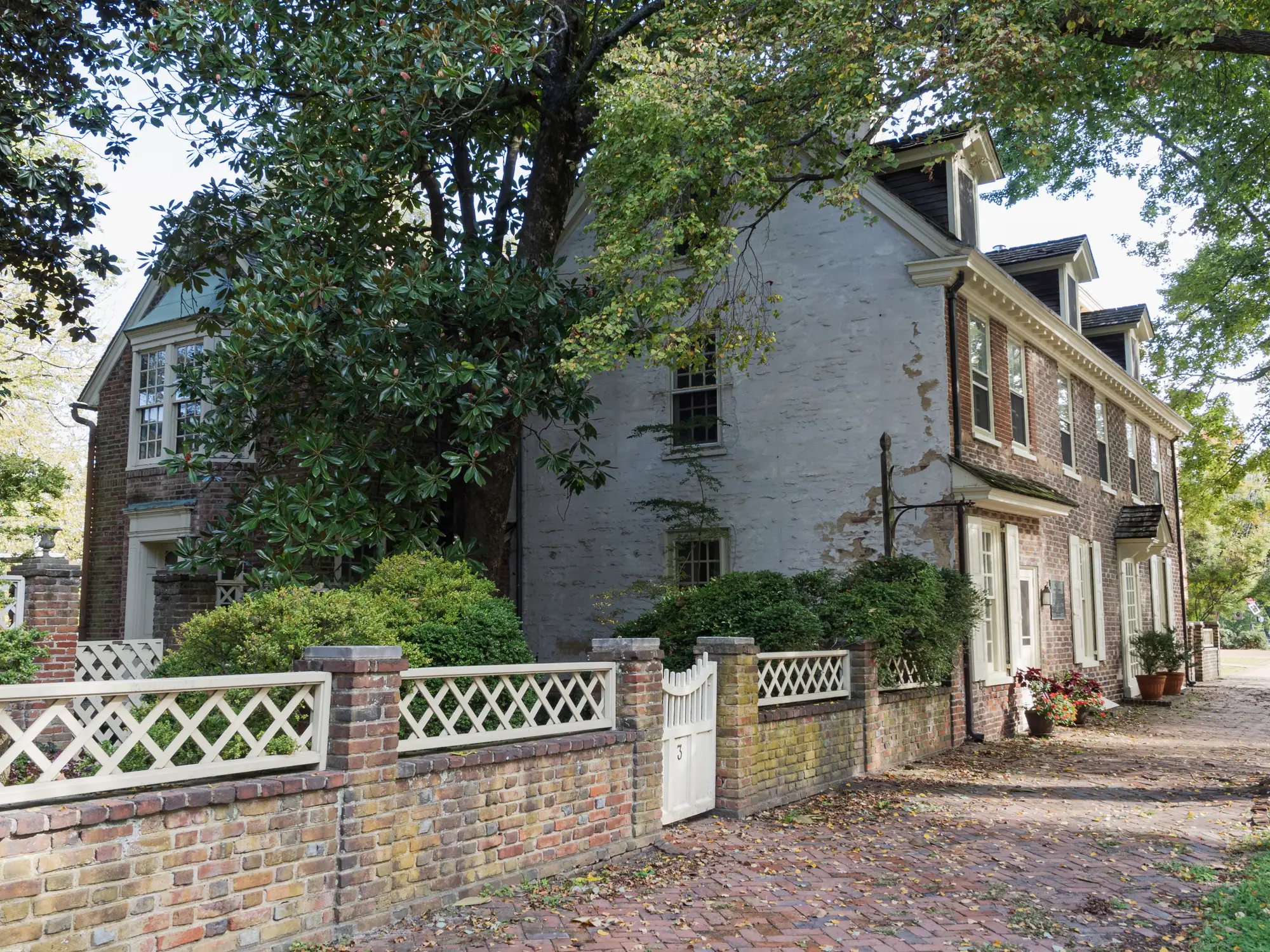 Historic brick house with white lattice fencing stands along a tree-shaded brick walkway.