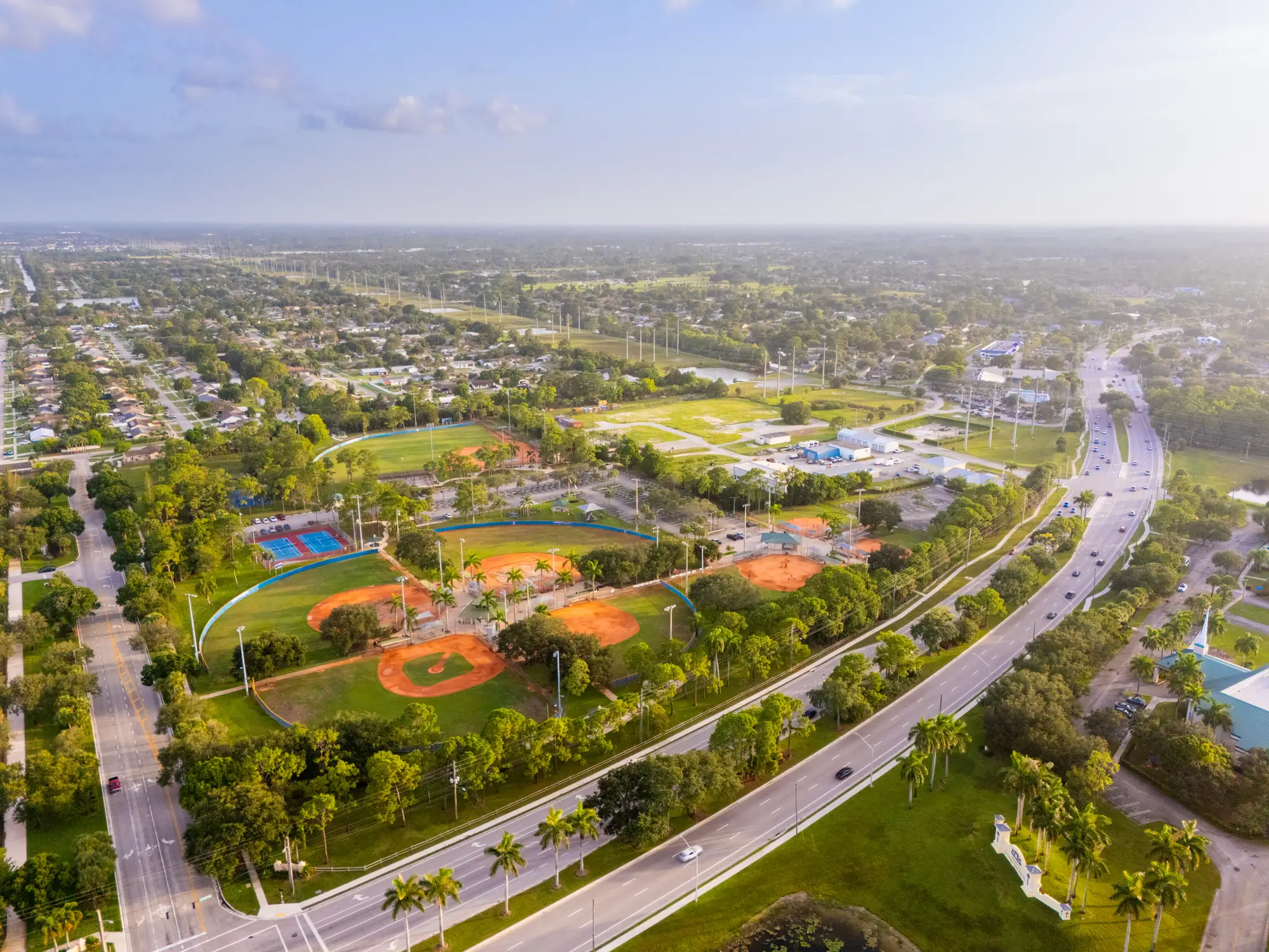 Aerial view of a suburban sports park with baseball fields, tennis courts, roads, trees, and surrounding neighborhoods.