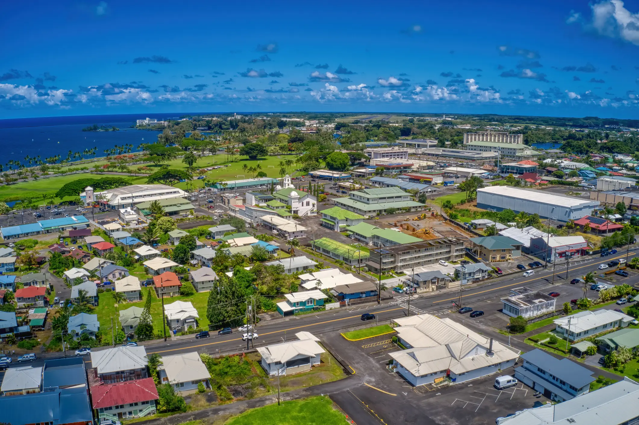 Aerial view of a coastal town with roads, buildings, green spaces, and a blue bay under scattered clouds.