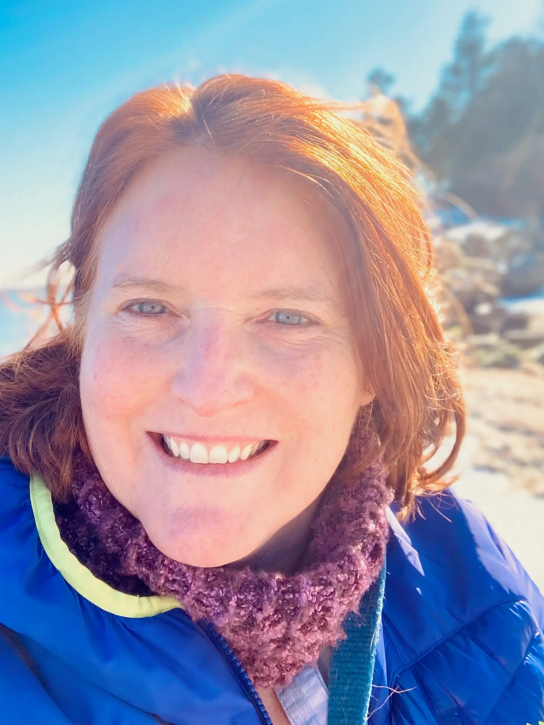 Smiling person in a blue jacket stands on a sunny beach with rocks, ocean, and trees behind them.