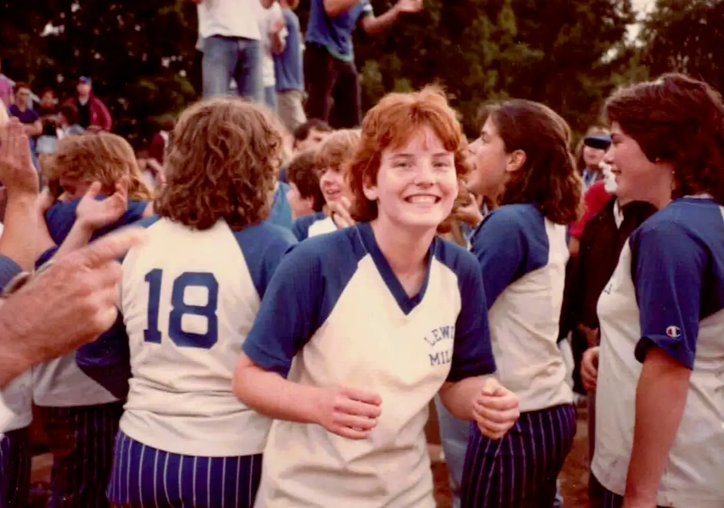 Uniformed players gather outdoors, with one smiling toward the camera amid teammates and a crowd.