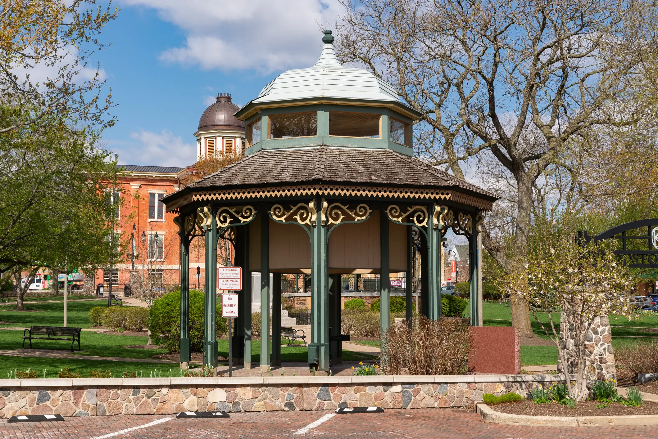 Ornate green gazebo stands in a landscaped park near a brick building with a domed tower.