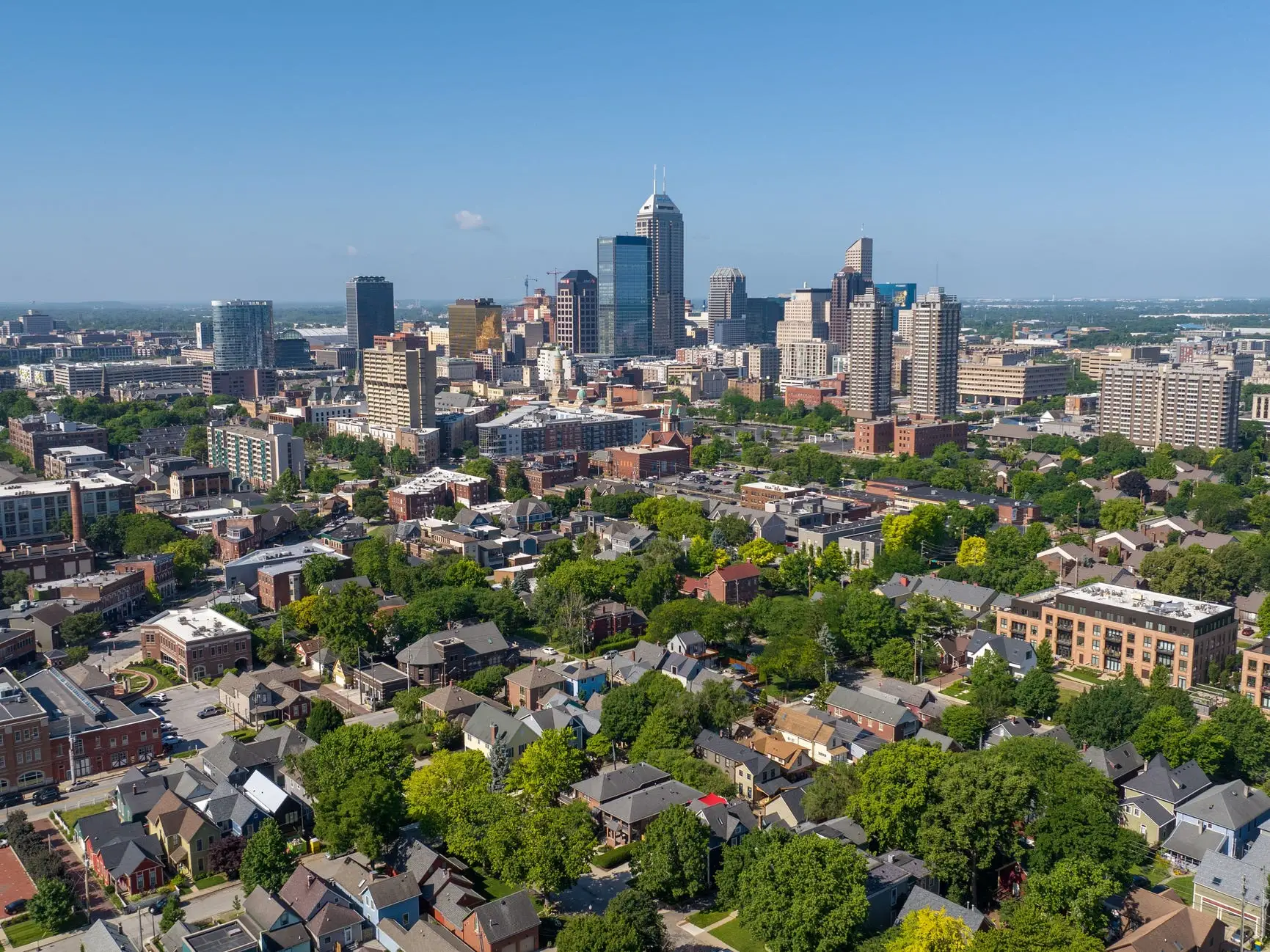 Aerial view of downtown Indianapolis skyline with surrounding residential neighborhoods and tree-lined streets under a clear blue sky.