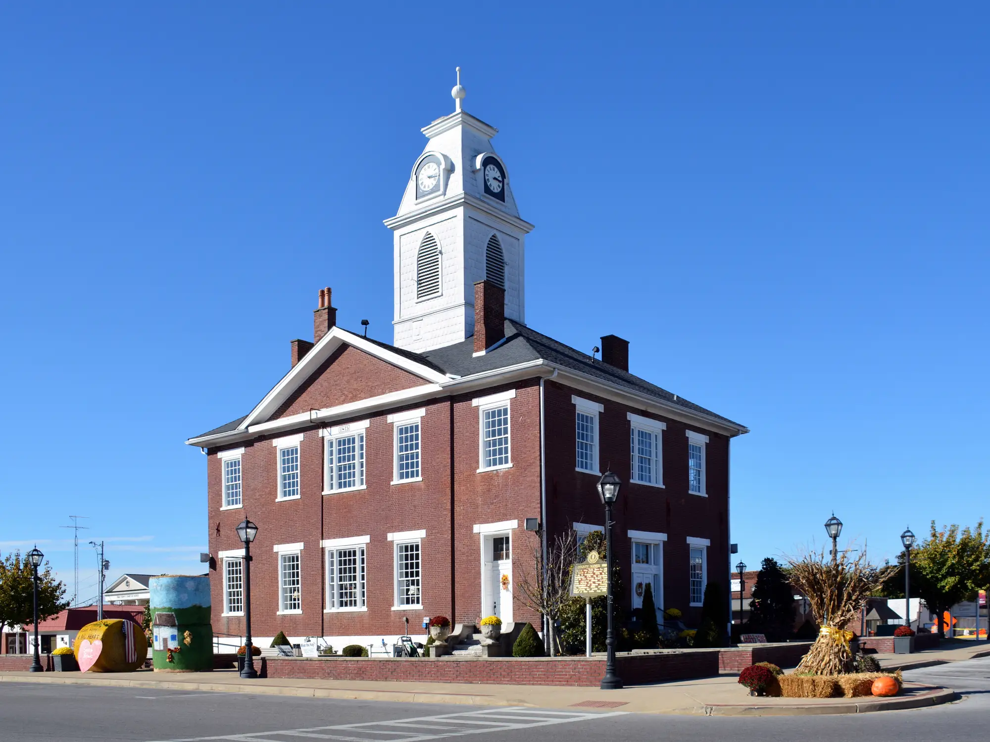 Red brick courthouse with white clock tower stands on a decorated town square under a clear blue sky.