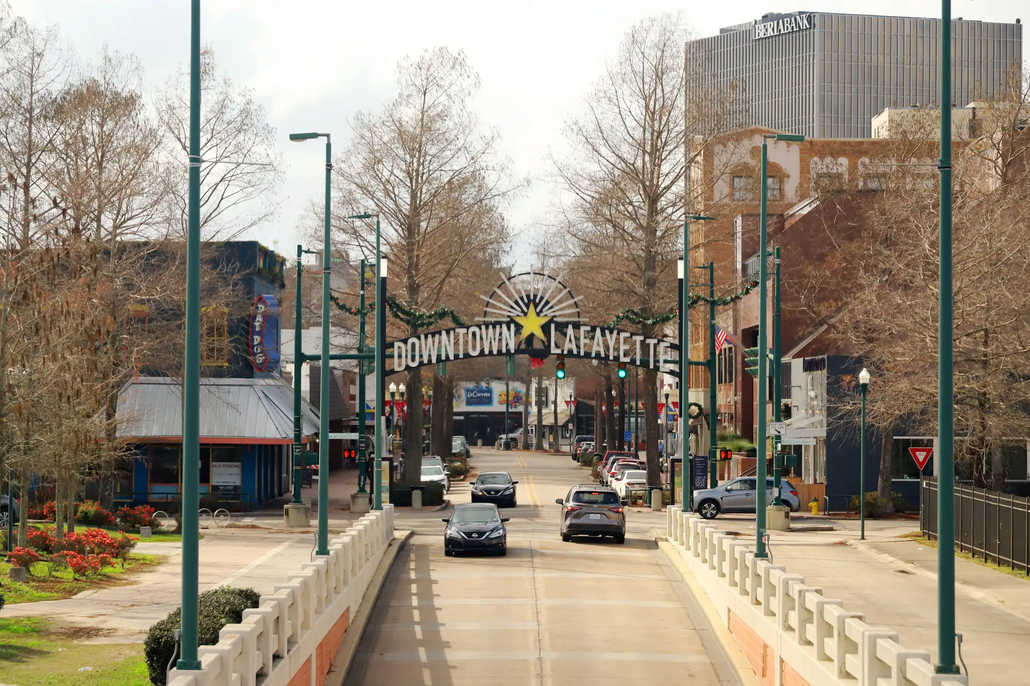 Cars drive under a DOWNTOWN LAFAYETTE arch on a city street lined with trees and storefronts.