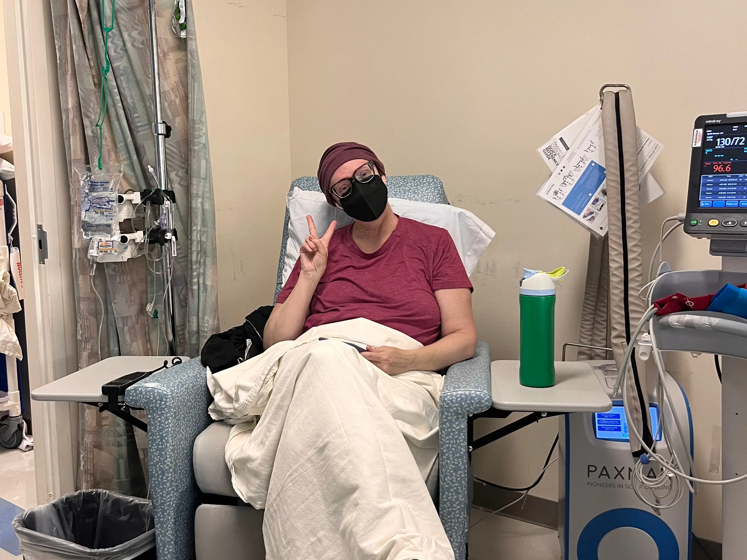 Person wearing a face mask sits in a medical infusion chair with blanket, IV equipment, and monitors nearby.