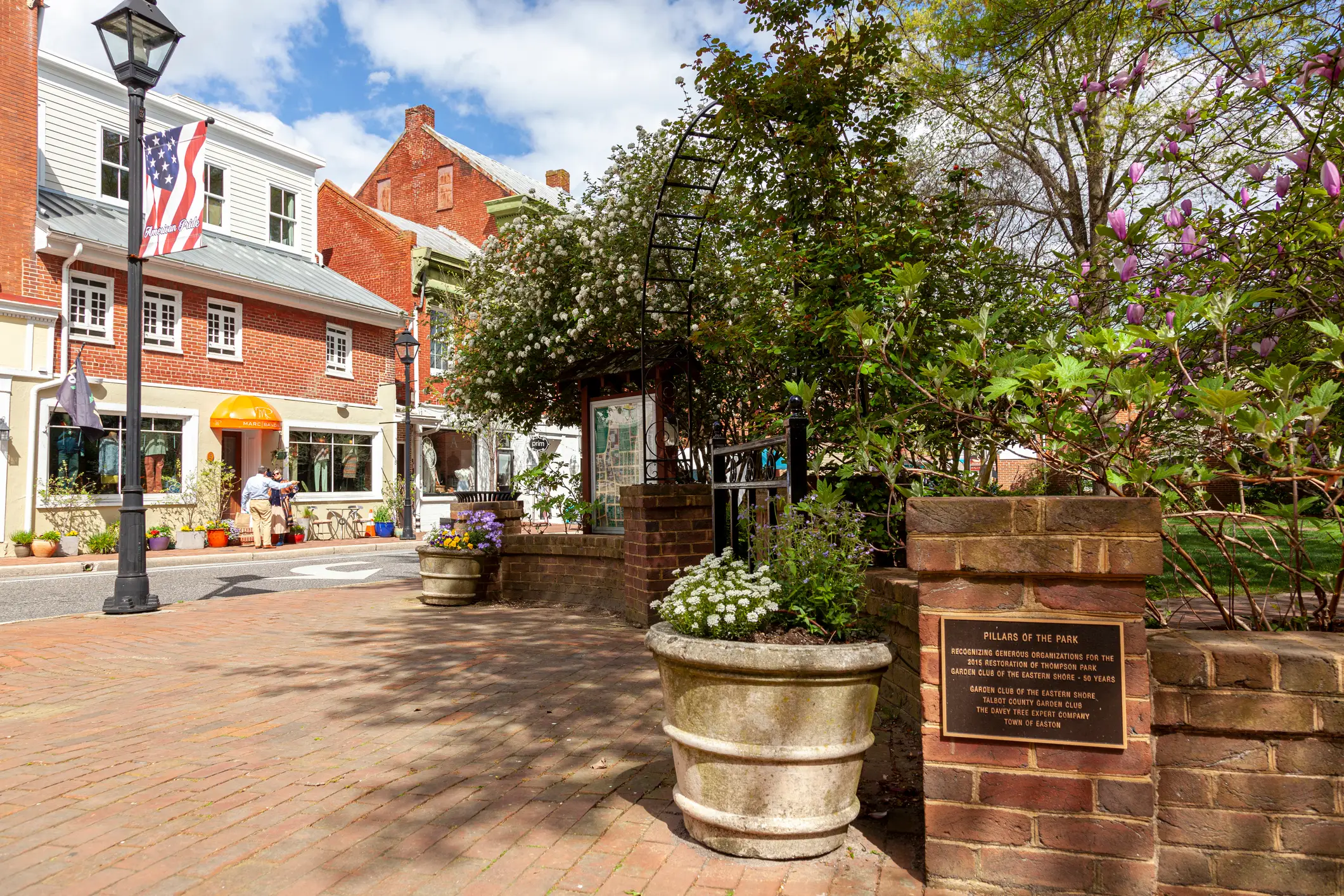 Brick sidewalk and garden entrance line a historic downtown street with storefronts, planters, and flowering trees.