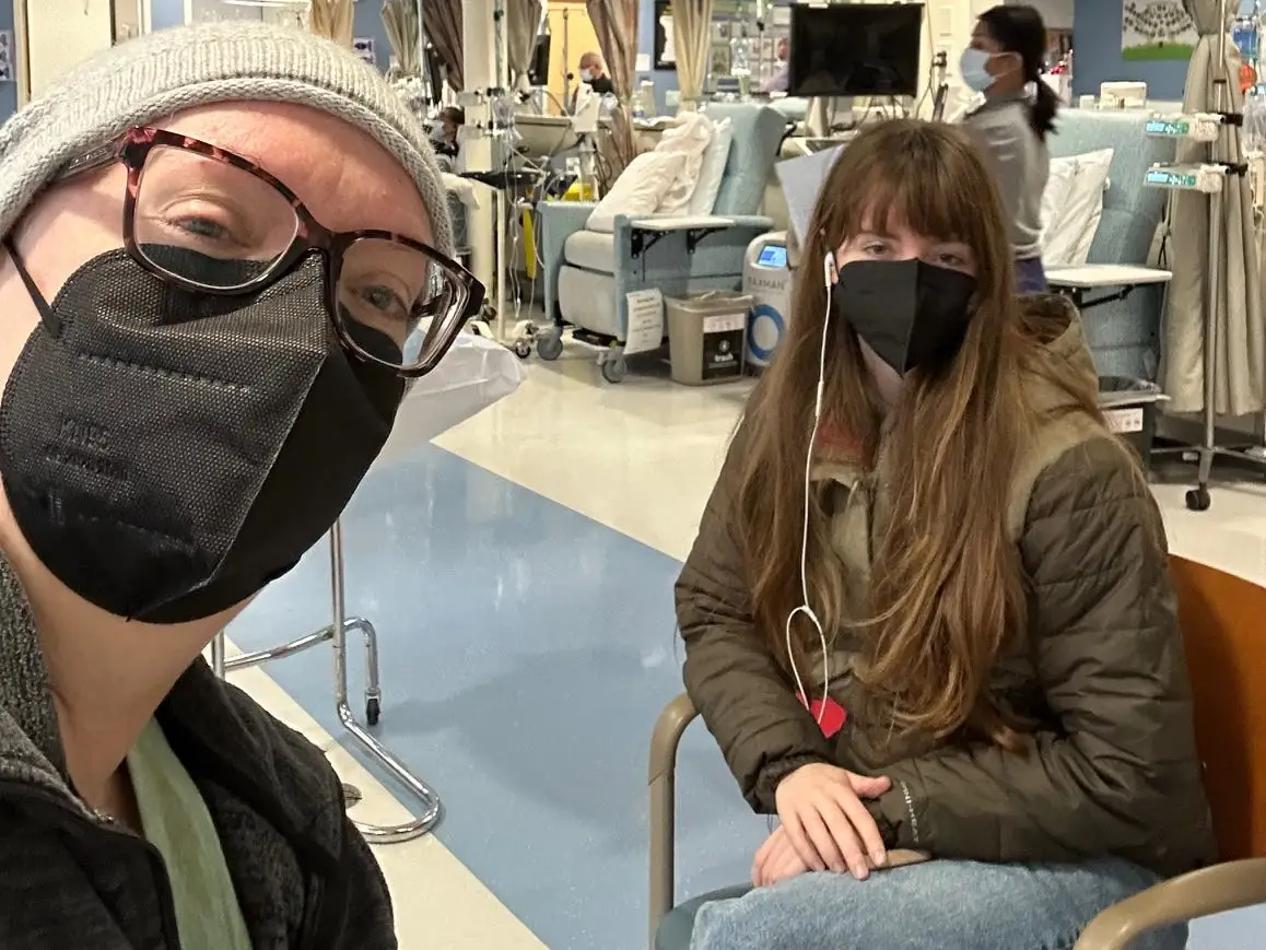 Two masked people sit in a clinical treatment room with infusion chairs and medical equipment in the background.
