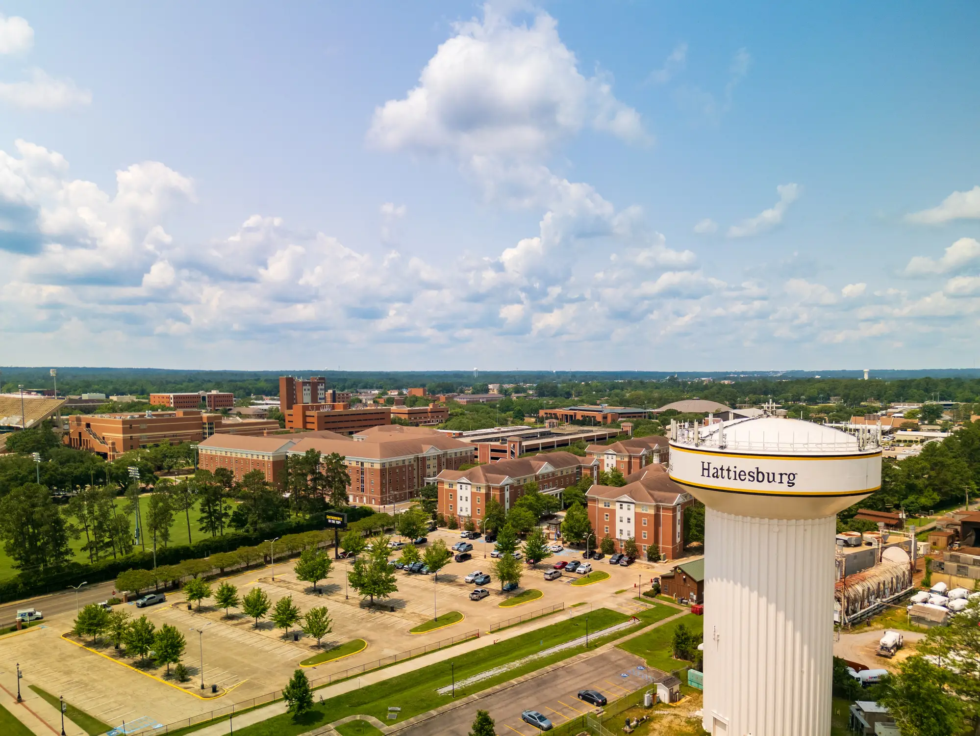 Aerial view of Hattiesburg water tower beside brick campus buildings, parking lots, trees, and a cloudy sky.