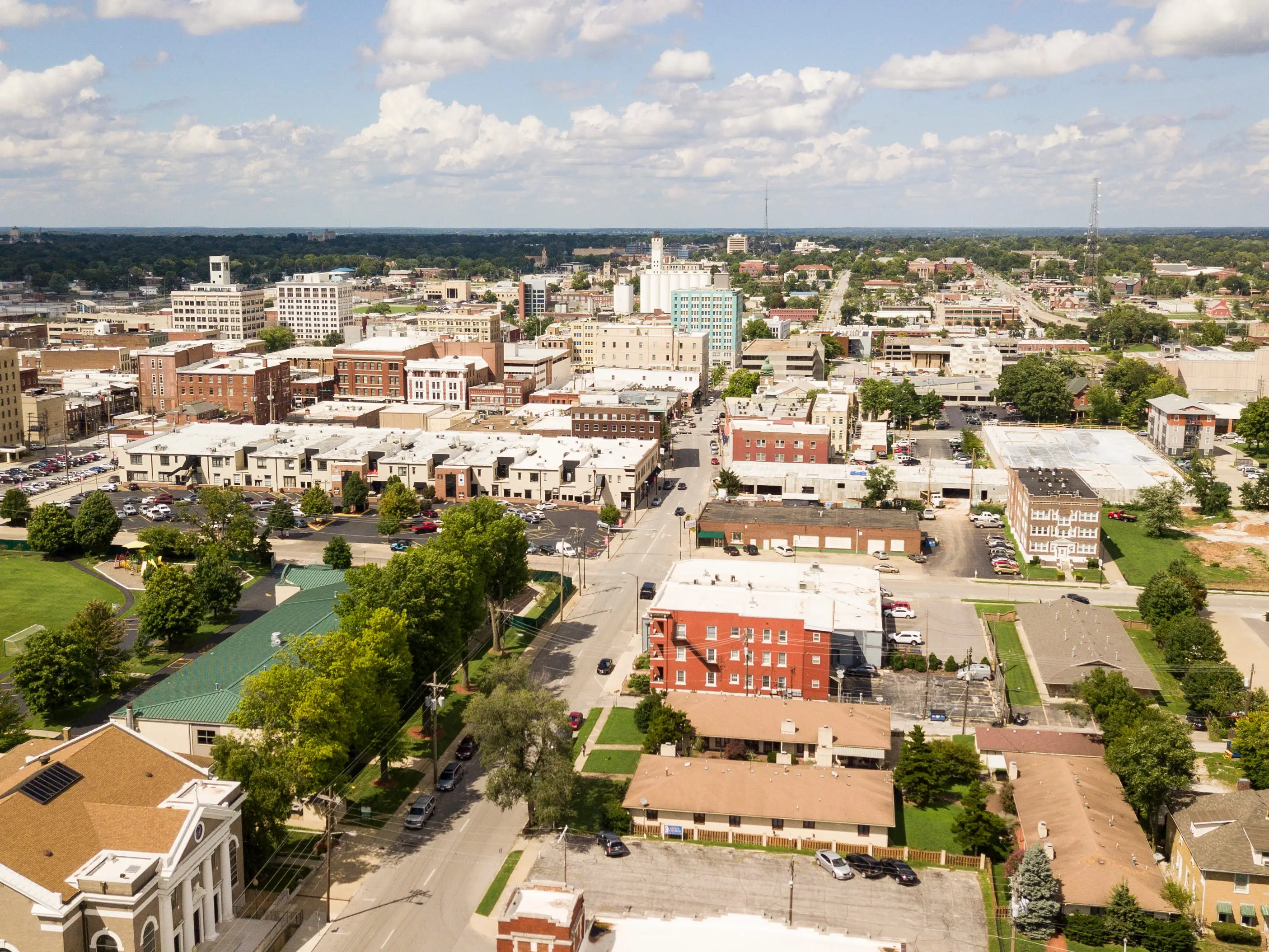 Aerial view of a downtown area with low-rise buildings, streets, parking lots, trees, and a partly cloudy sky.