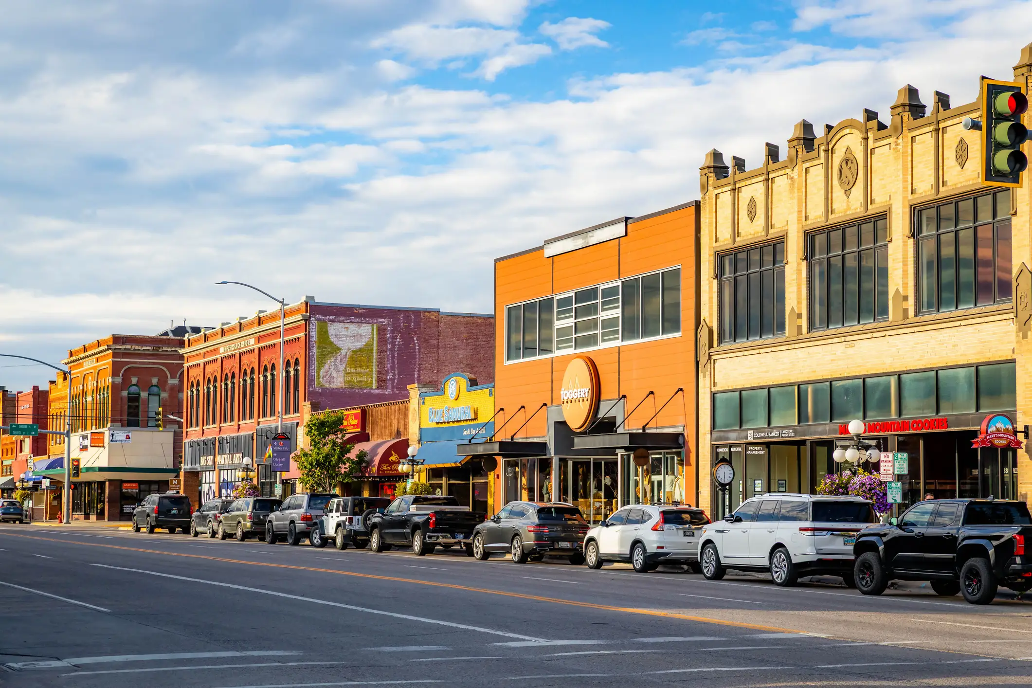 Downtown commercial street with historic storefronts, parked cars, and a traffic light under a partly cloudy sky.