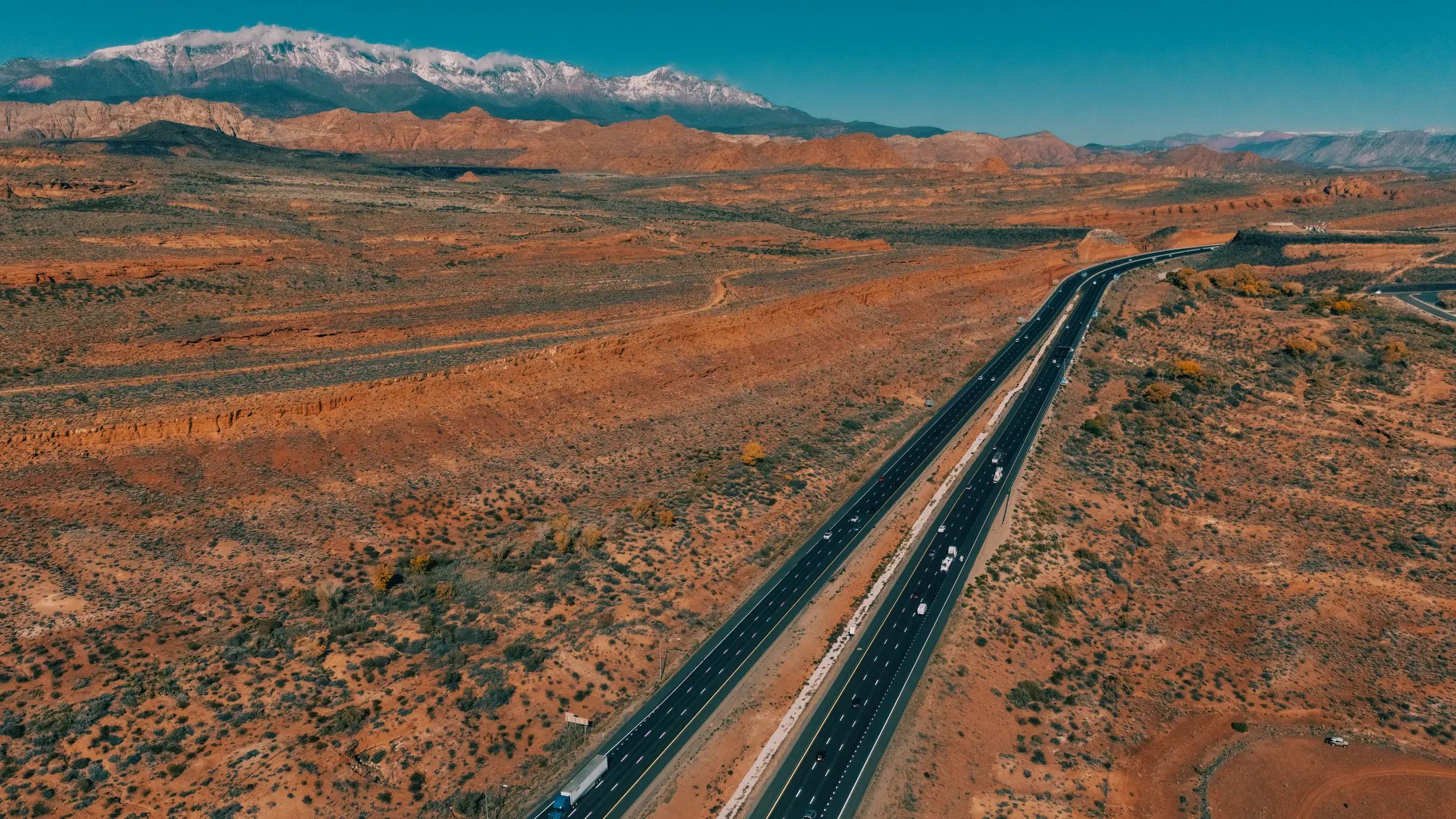 Aerial view of a divided highway crossing red desert terrain toward snow-capped mountains under a clear sky.
