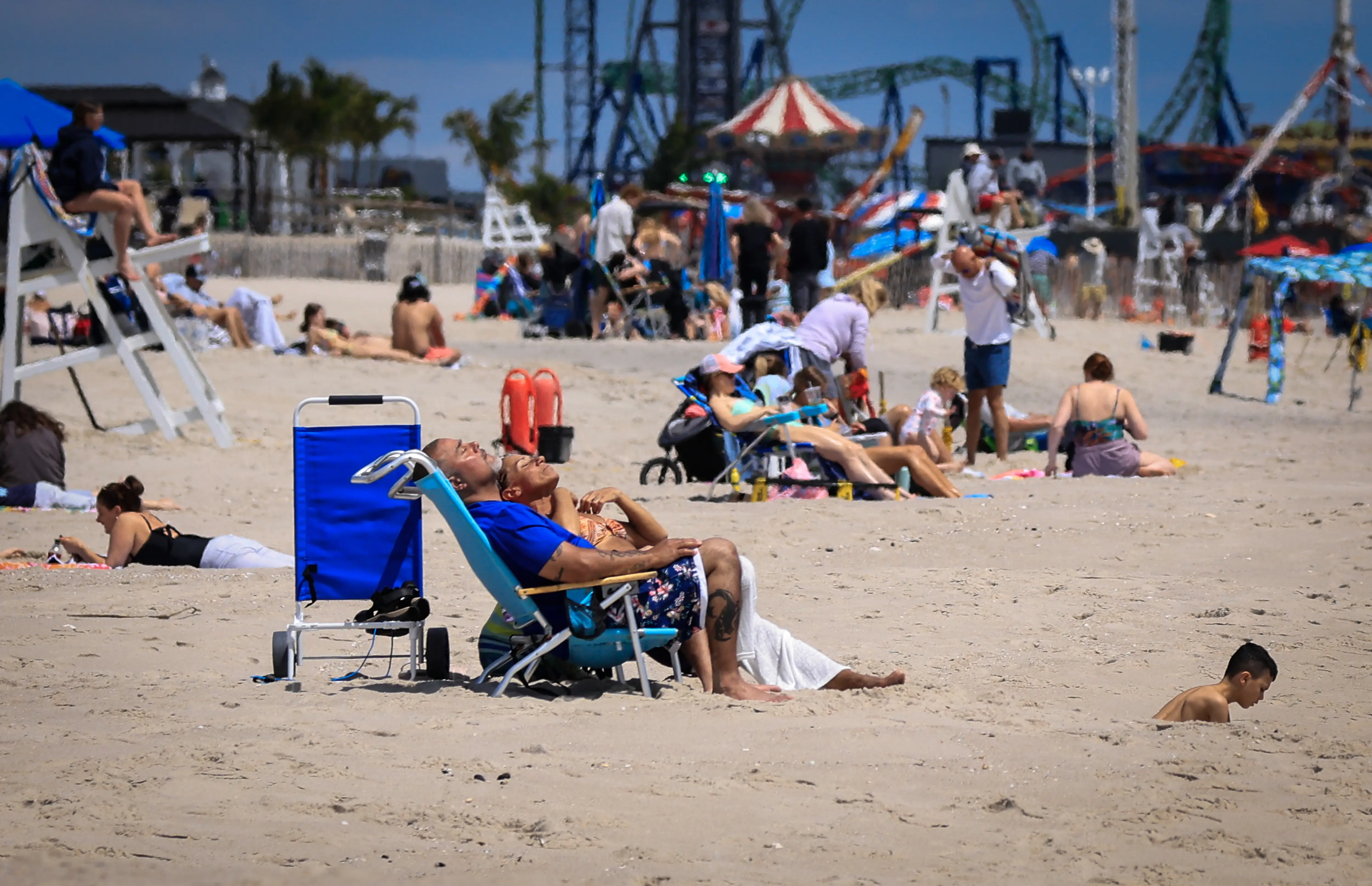 Beachgoers relax on a sandy beach with a lifeguard chair and amusement park rides in the background.