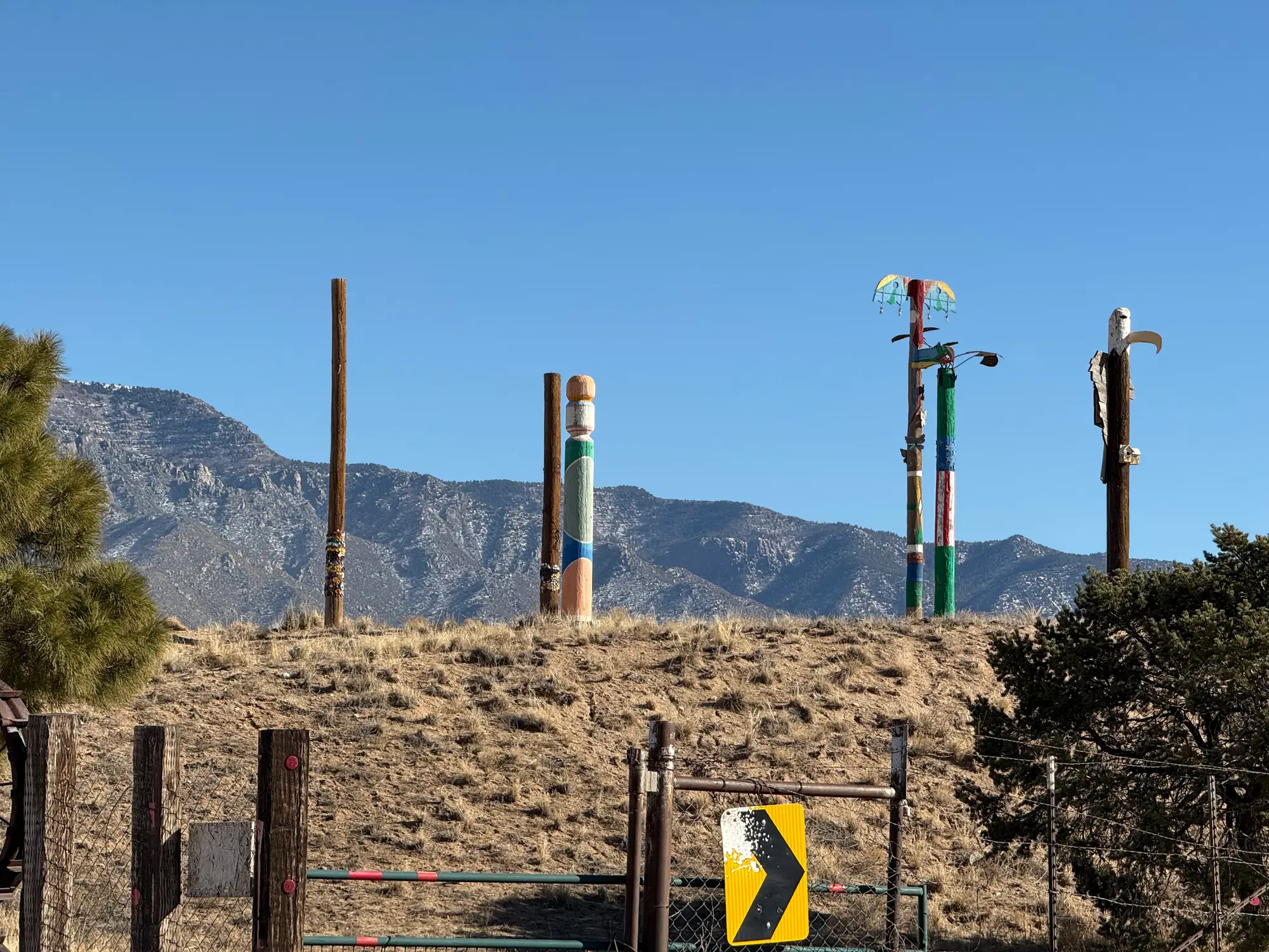 Colorful pole sculptures stand on a dry hillside with mountains and blue sky in the background.