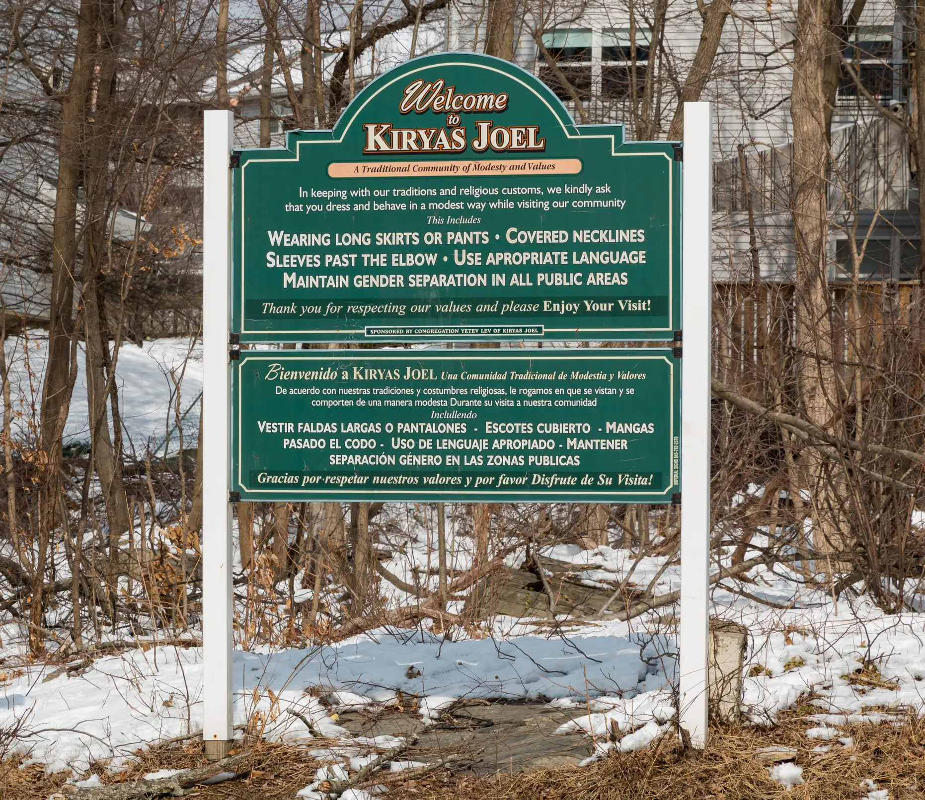 Welcome to Kiryas Joel sign displays community dress and behavior guidelines in English and Spanish beside snowy woods.