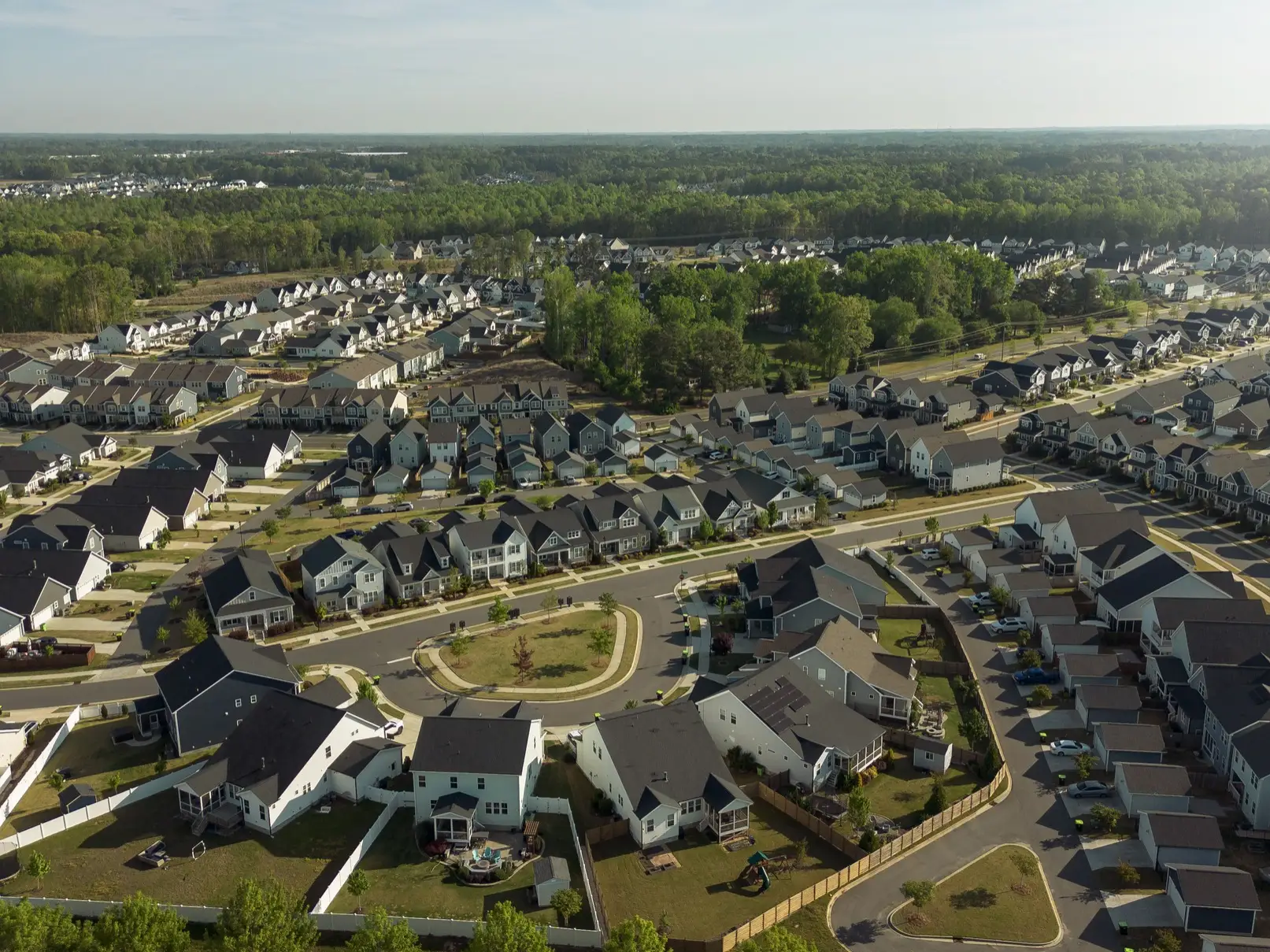 Aerial view of a suburban housing development with rows of homes, streets, lawns, and surrounding trees.