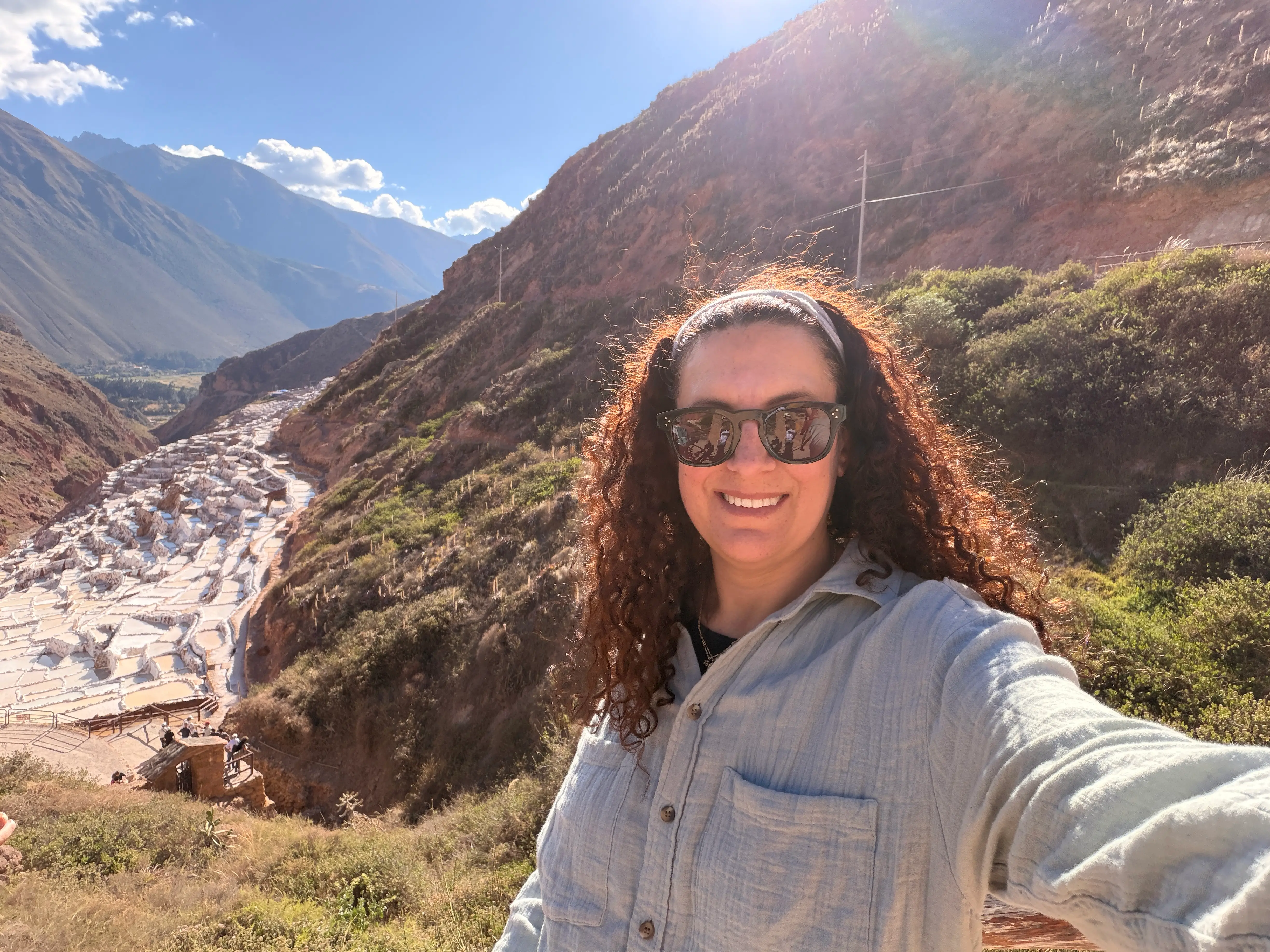 A smiling traveler takes a selfie above terraced salt ponds in a sunlit mountain valley.