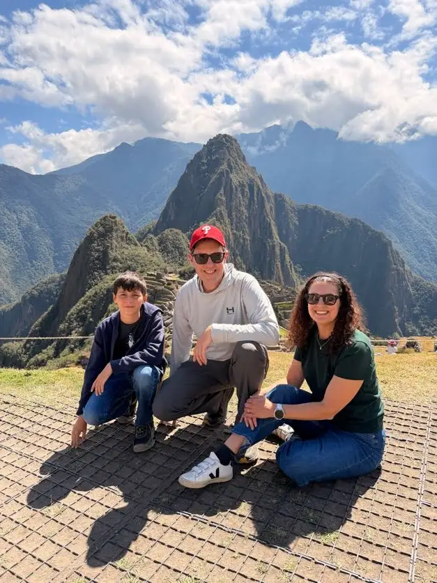 Three people pose on a stone terrace with Machu Picchu and surrounding mountains in the background.