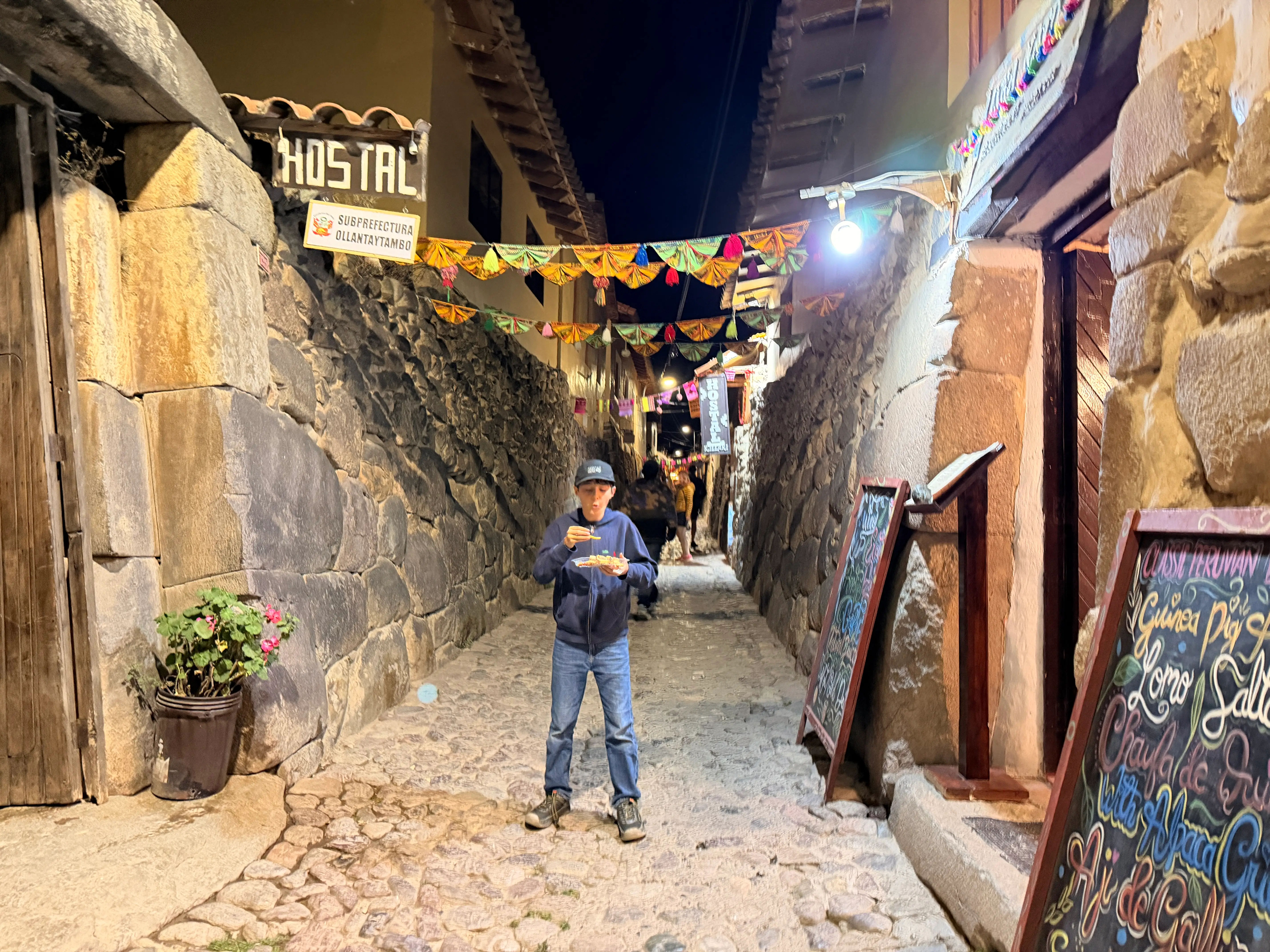 A person eats while standing in a lit cobblestone alley with stone walls, bunting, and hostal signage at night.