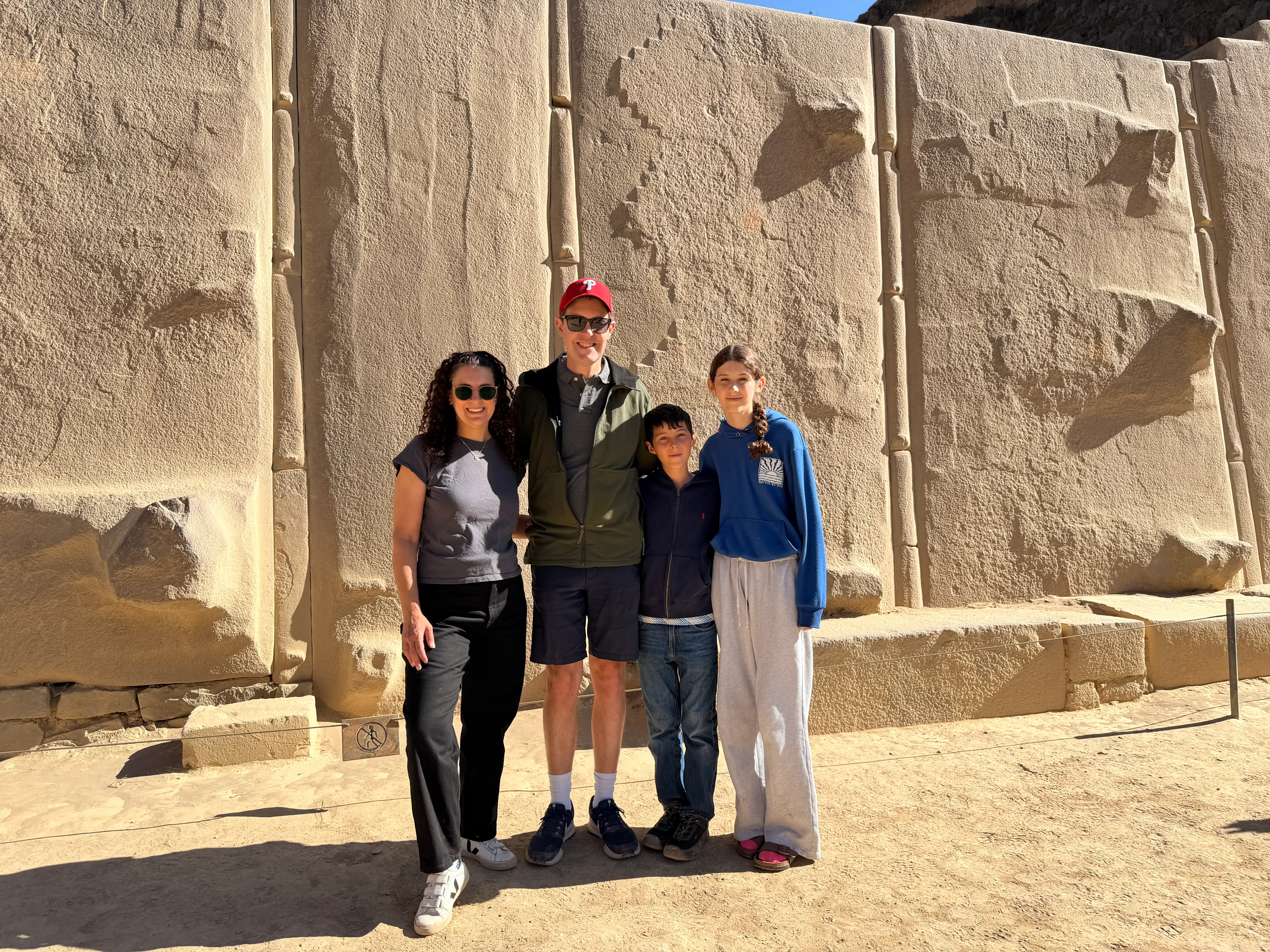 Four visitors pose in front of a large sunlit carved stone wall at an archaeological site.