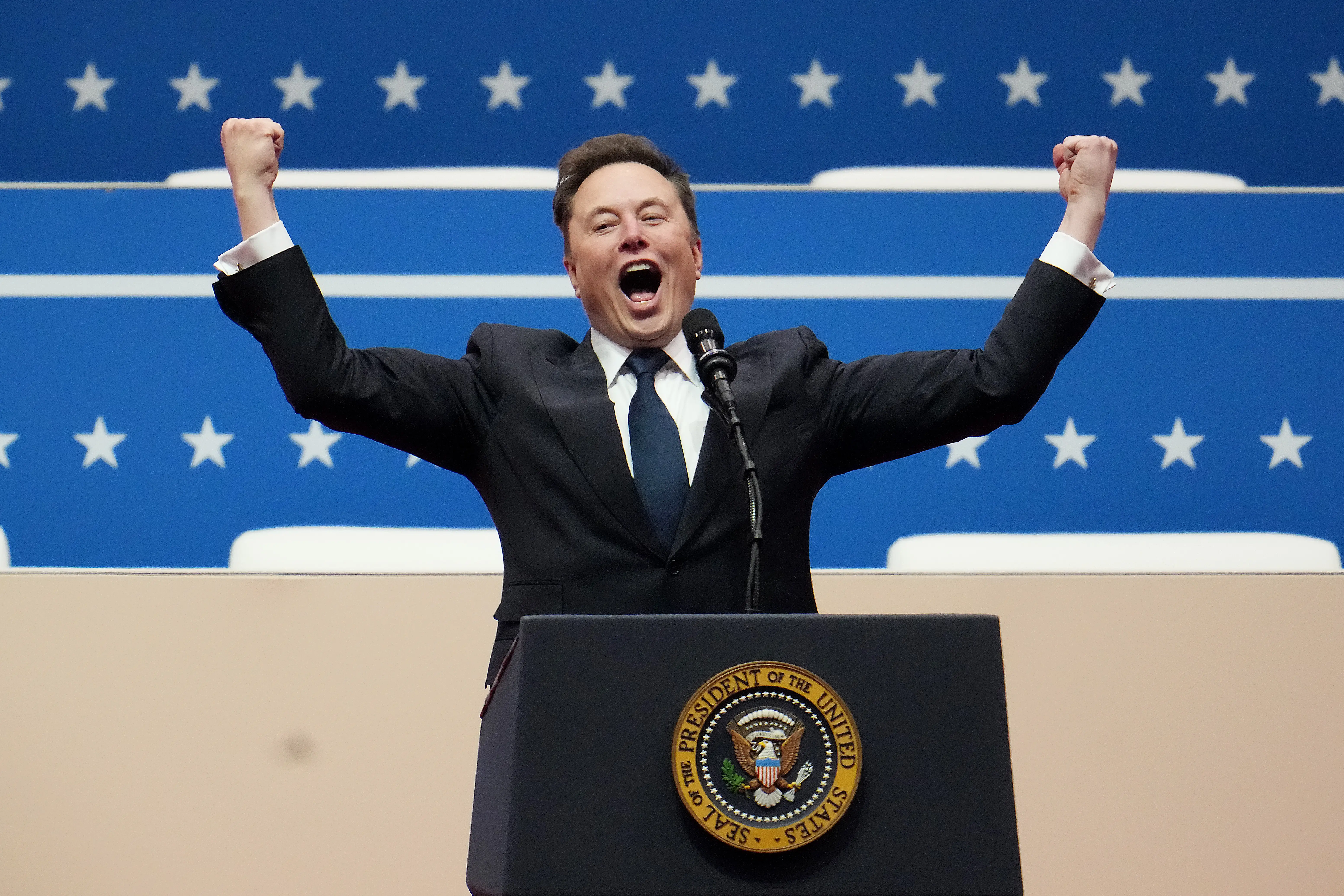 Elon Musk at a podium with arms stretched in the air