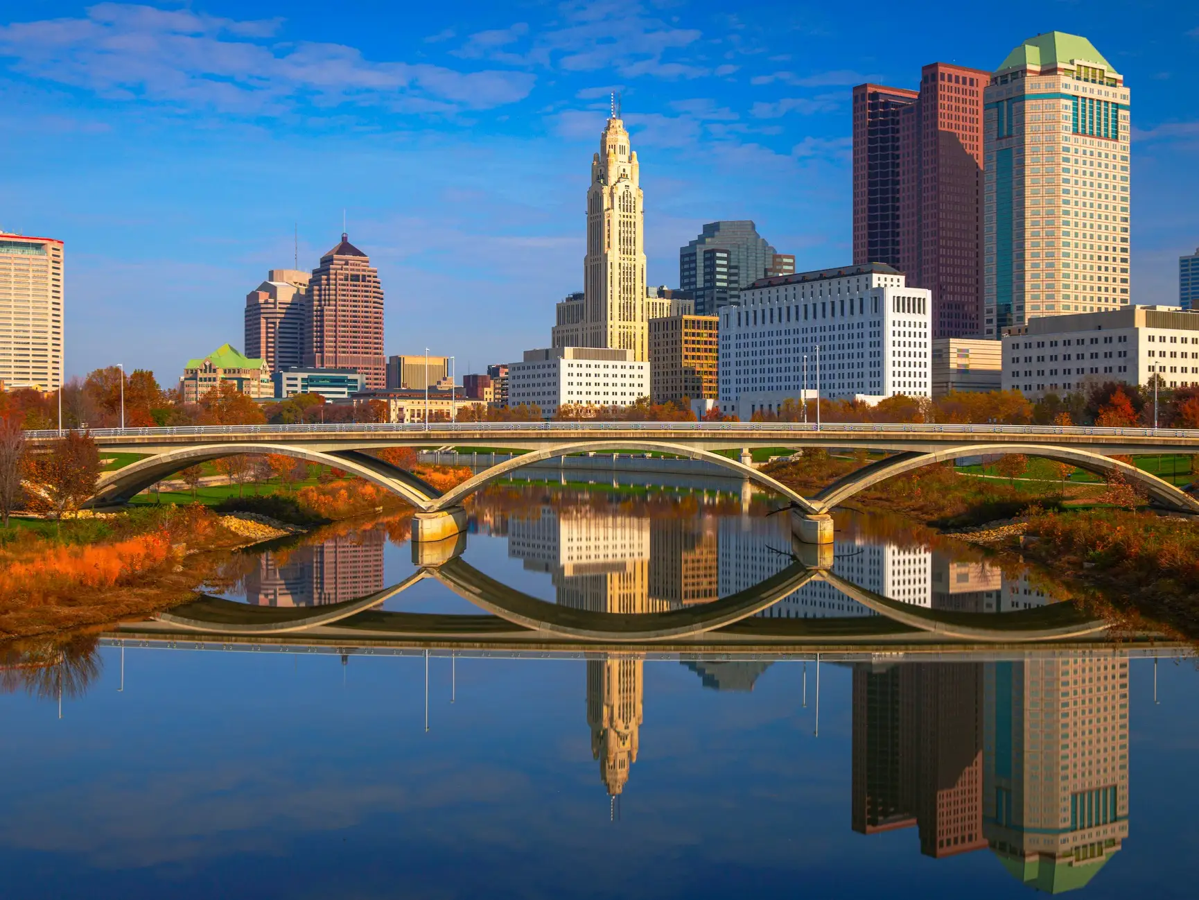 Downtown Columbus skyline and arched bridge reflected in the Scioto River under a blue sky.