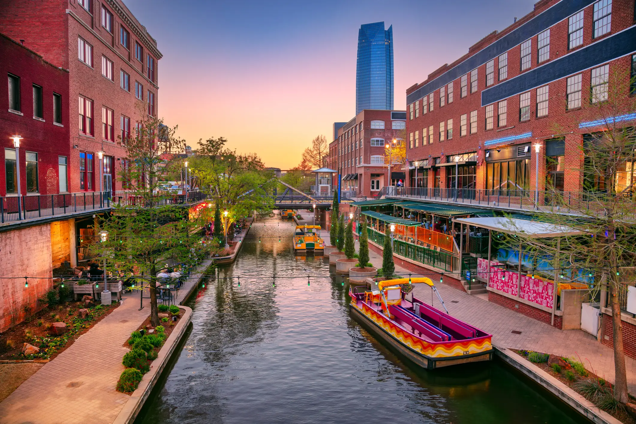 Brick-lined canal district at sunset with water taxis, walkways, restaurants, and a tall glass tower in the background.