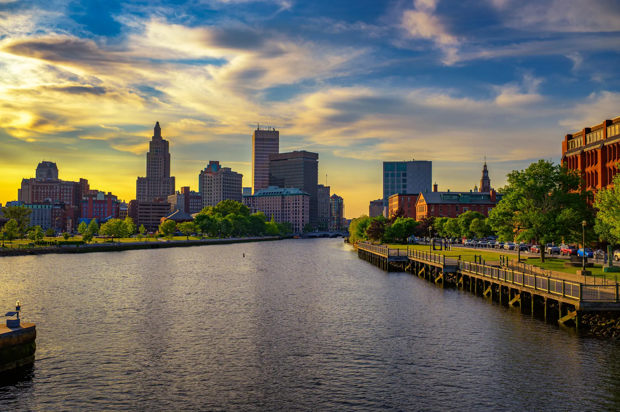 Providence skyline and riverfront at sunset with downtown towers, trees, boardwalk, and dramatic clouds.