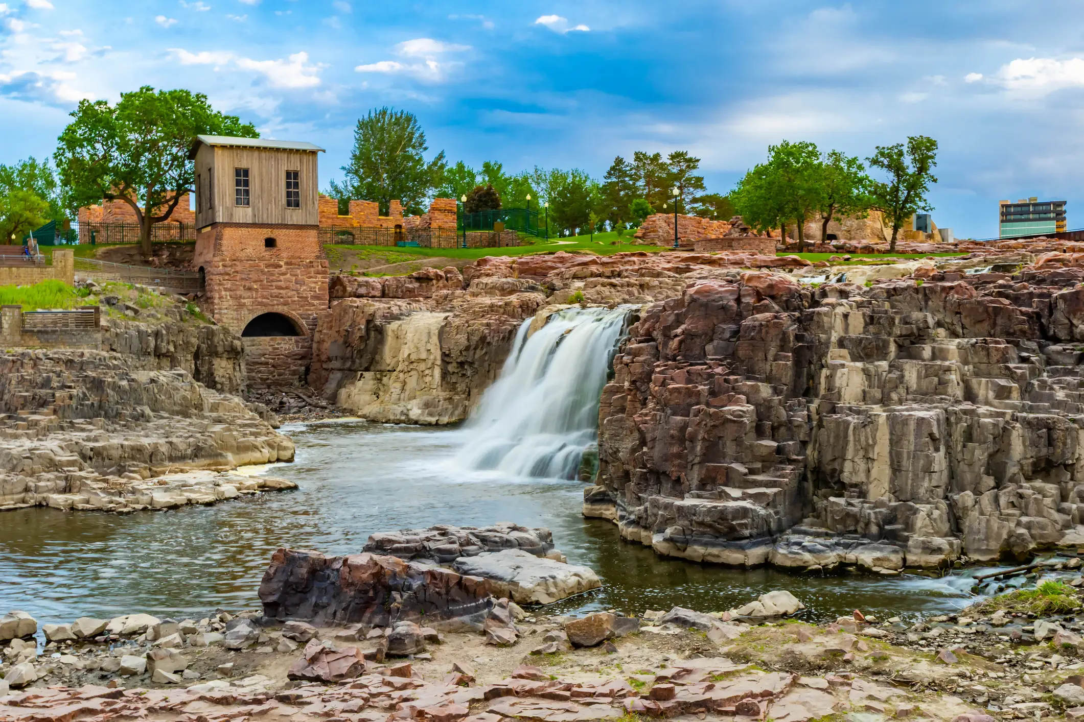 Waterfall flows through rocky cliffs beside historic stone structures and trees under a cloudy blue sky.