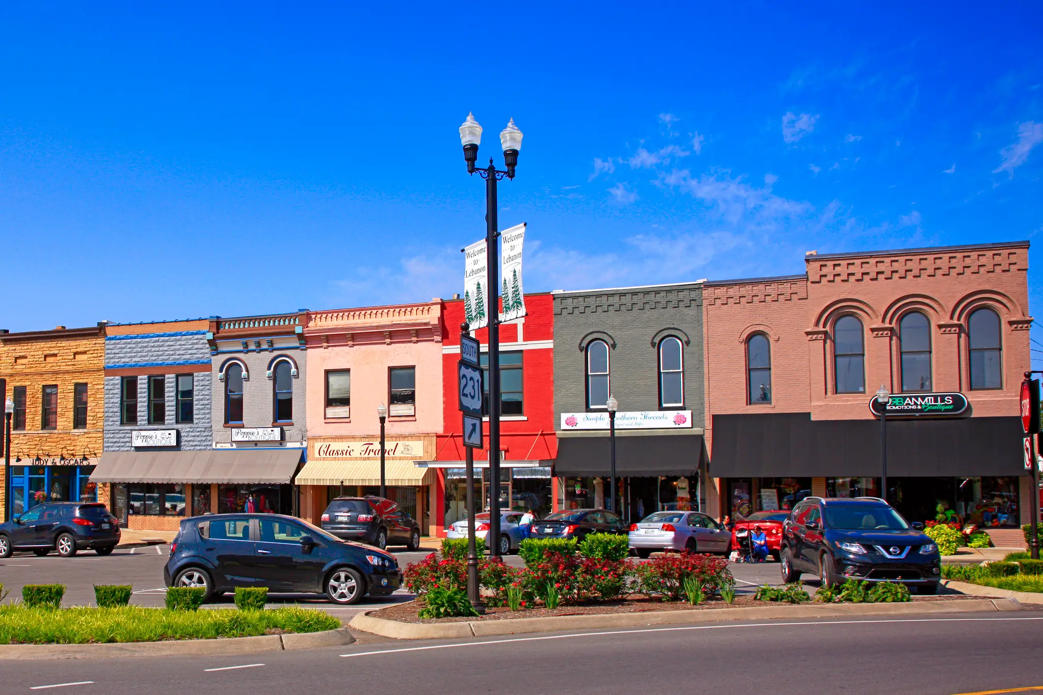Colorful brick storefronts, parked cars, a Route 231 sign, and flower beds line a sunny downtown street.
