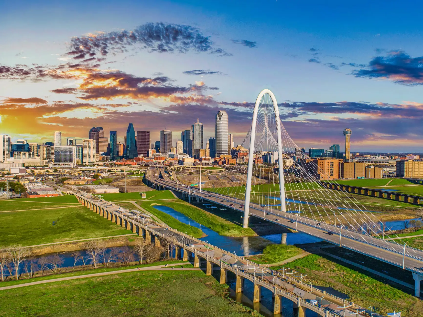 Dallas skyline at sunset with the Margaret Hunt Hill Bridge spanning green parkland and water in the foreground.