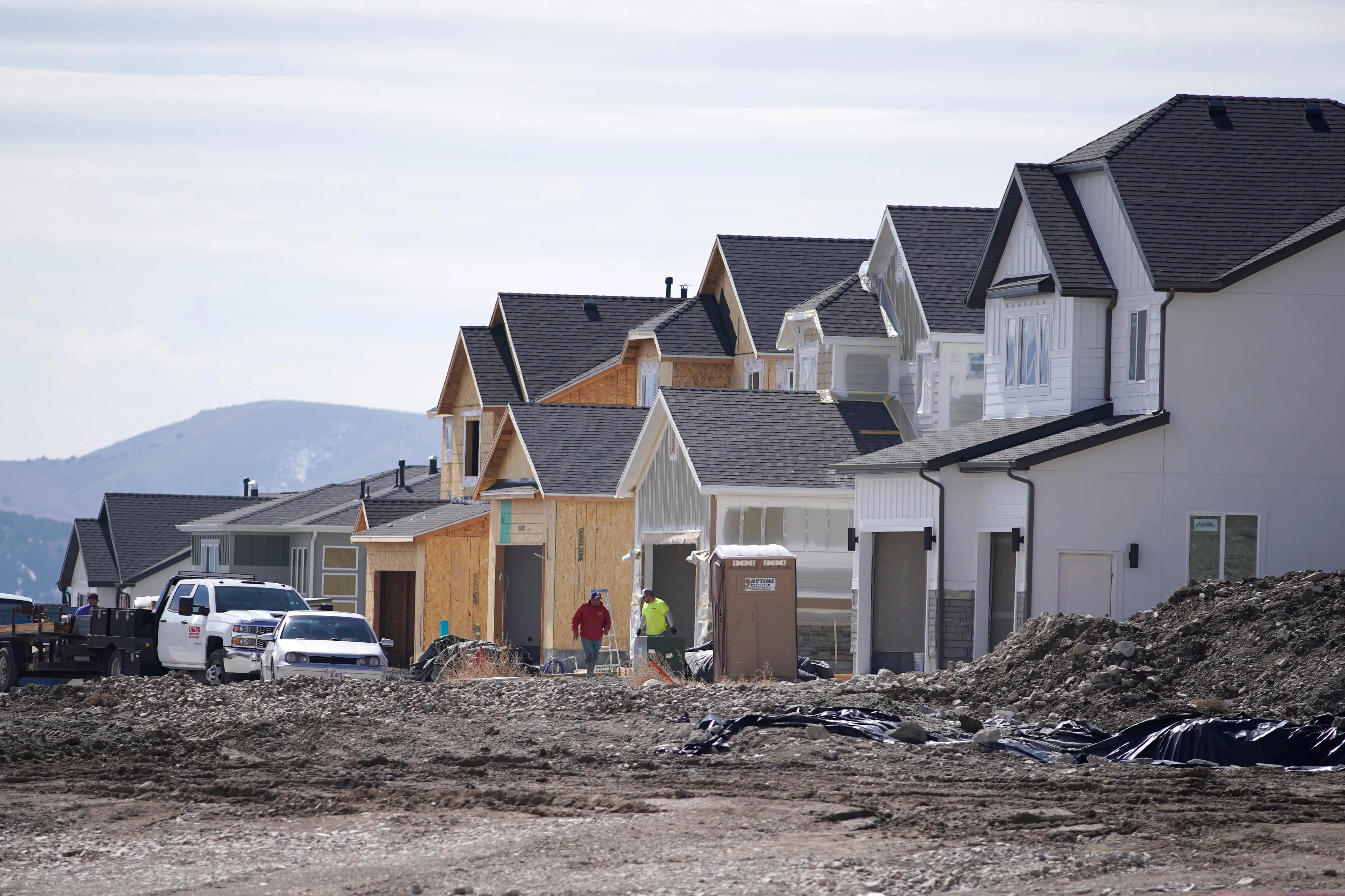 New houses under construction line a muddy residential development site with vehicles, workers, and mountains in the background.