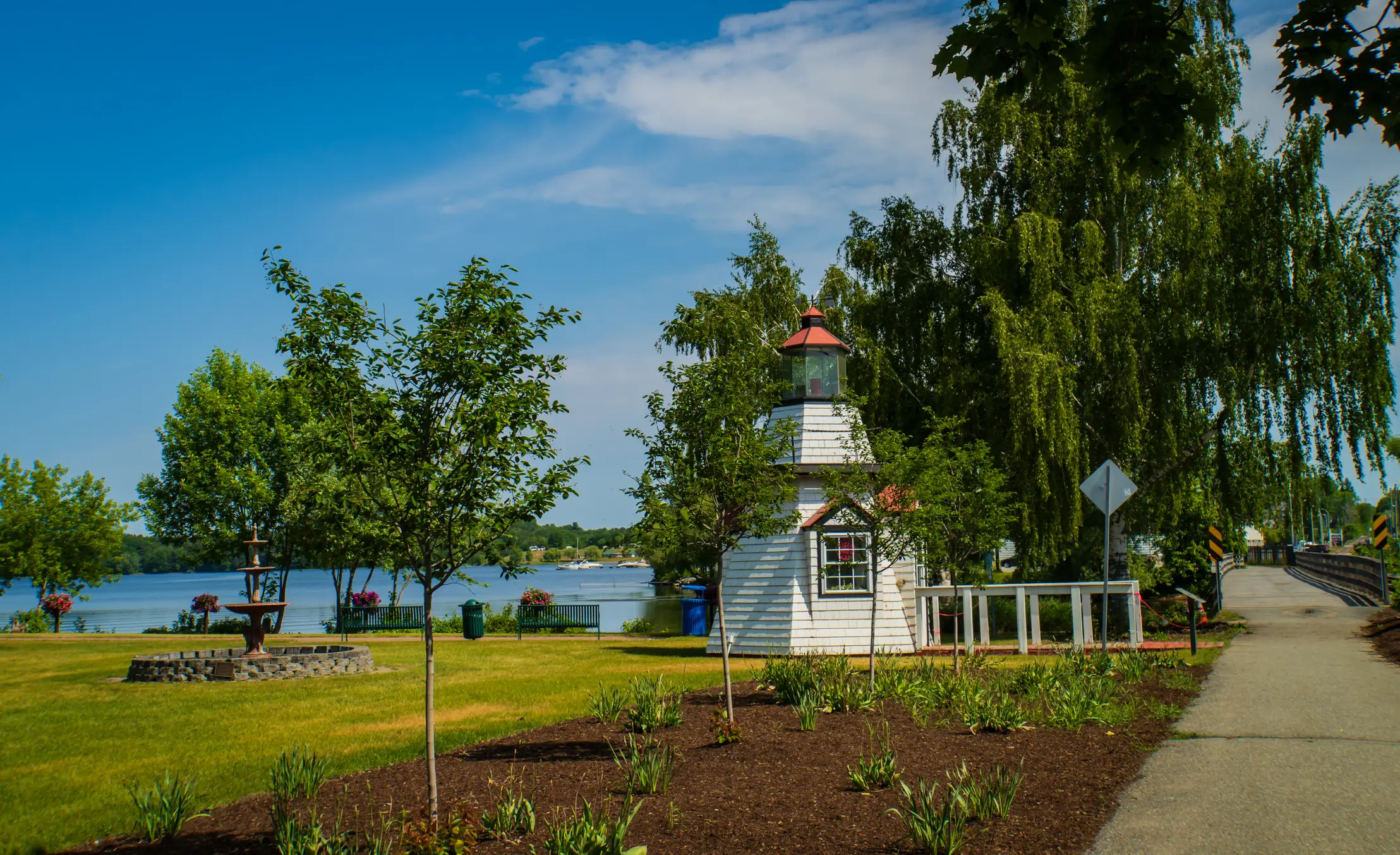 Small white lighthouse stands in a landscaped waterfront park beside a path, trees, and calm blue water.