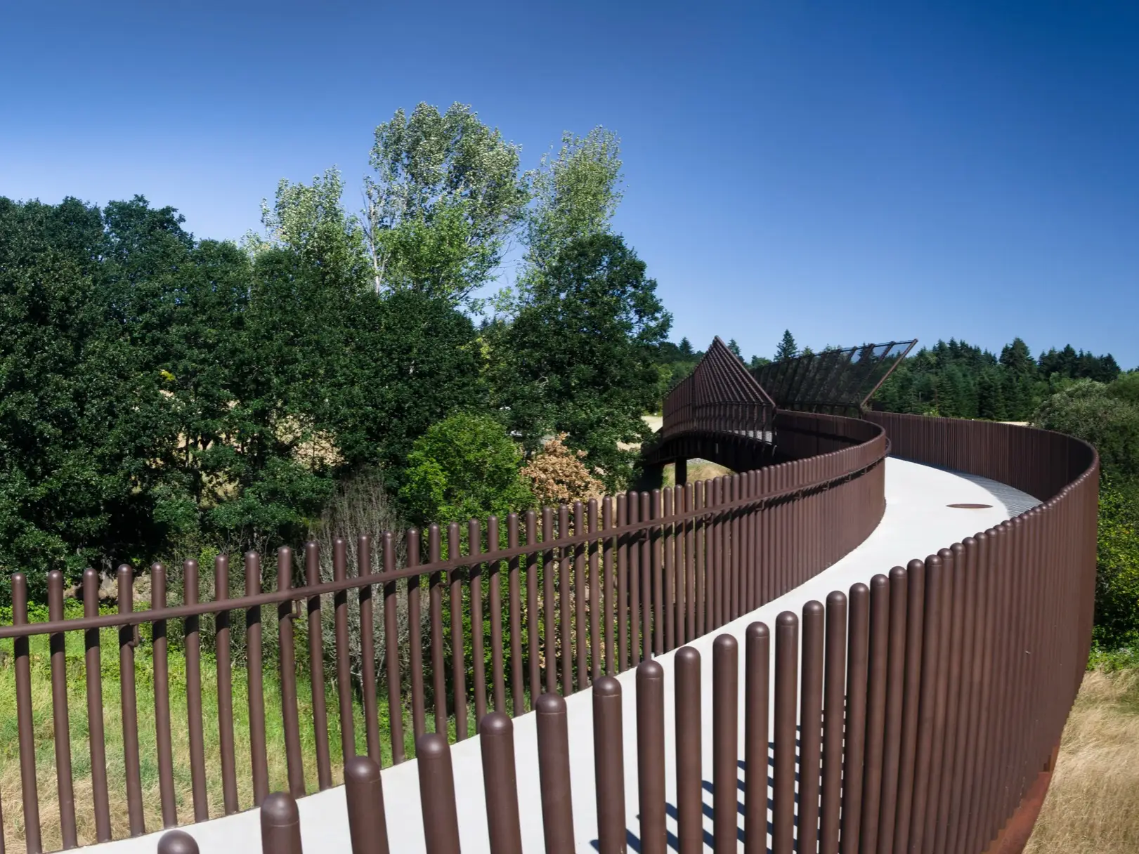Curved elevated walkway with brown railings winds through green trees under a clear blue sky.