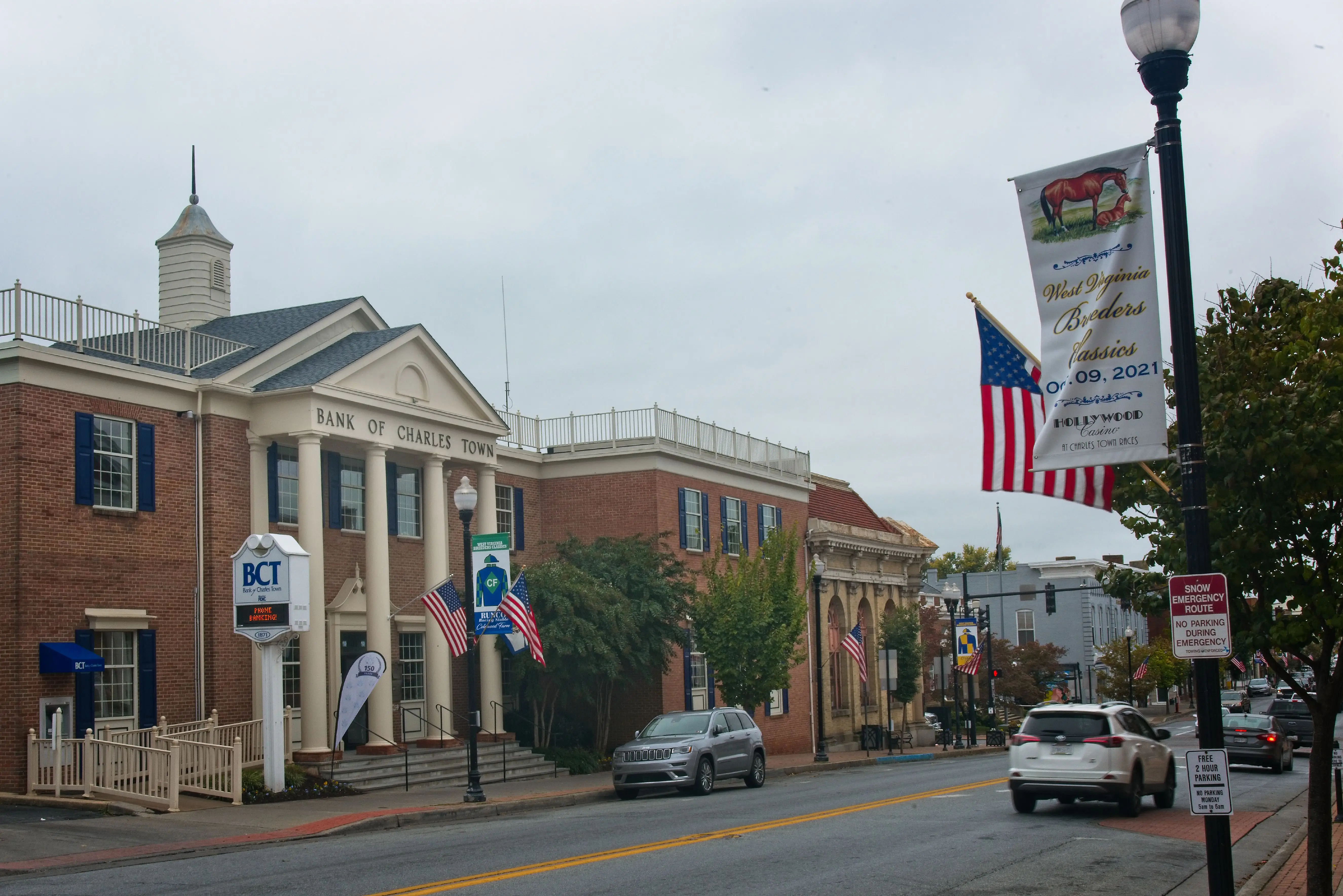 Downtown street with Bank of Charles Town building, American flags, parked cars, and lamppost banners under an overcast sky.