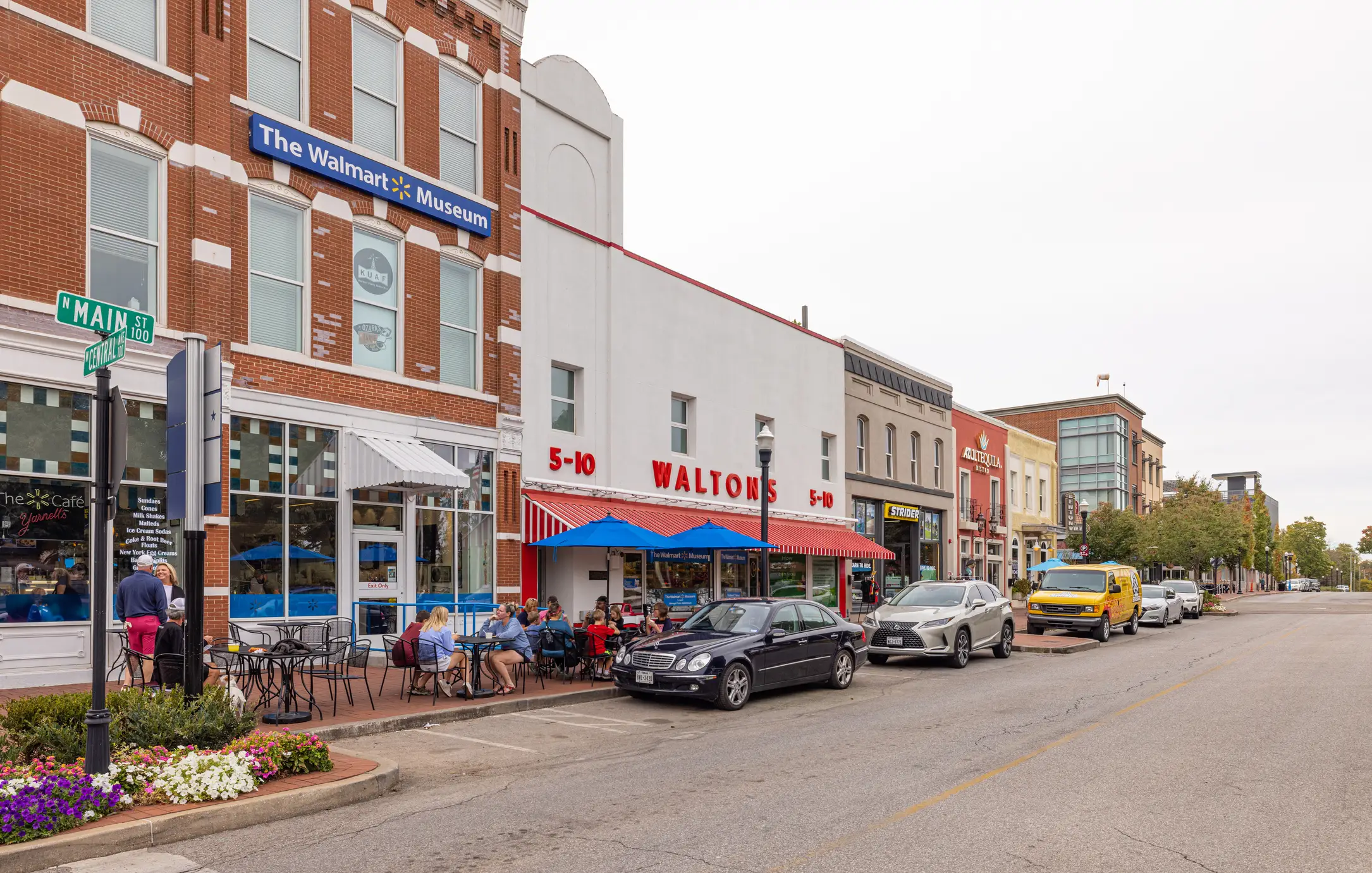 Street view of The Walmart Museum and Walton's 5-10 storefronts with outdoor seating and parked vehicles.