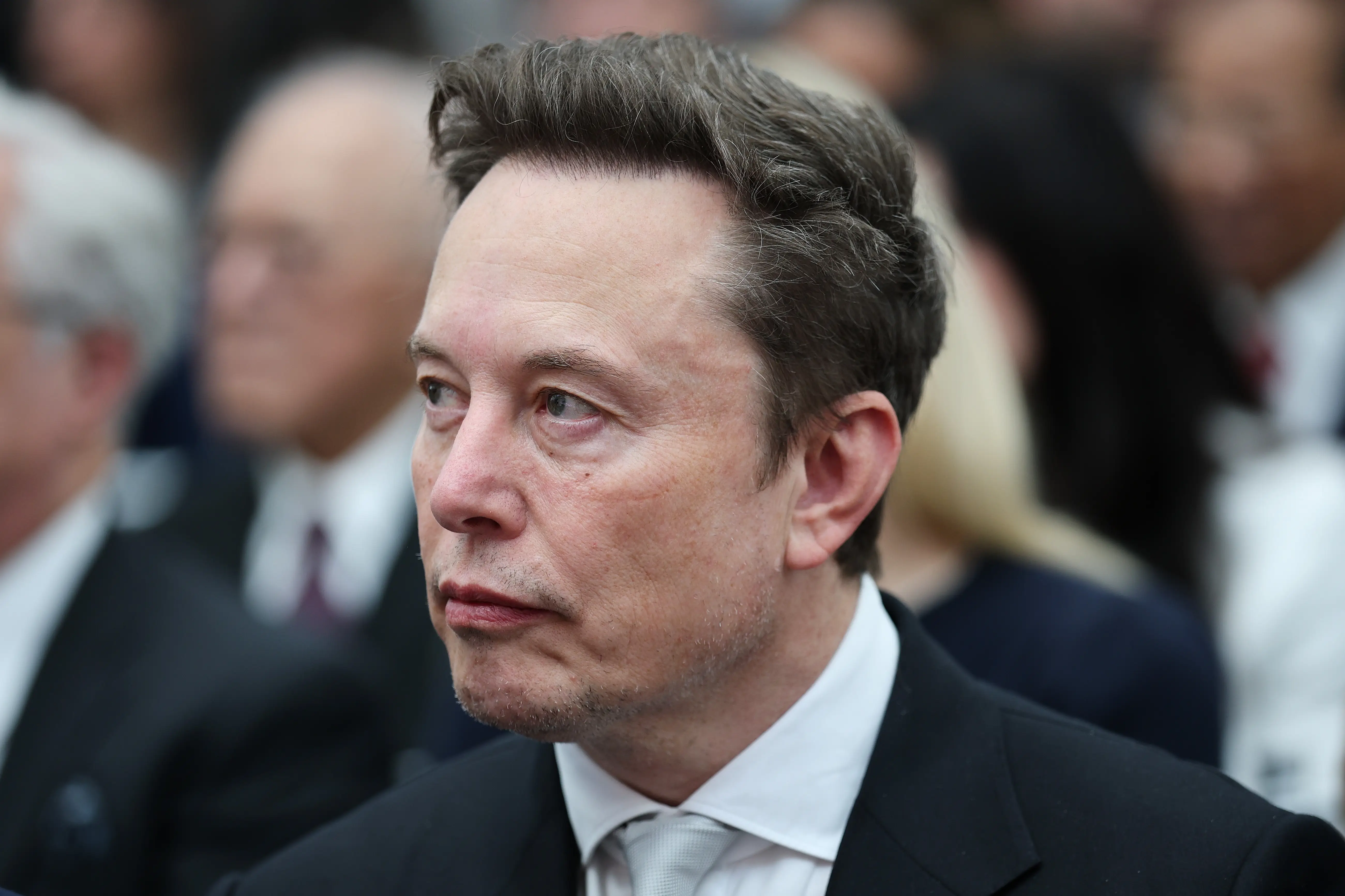Elon Musk sits in a formal setting wearing a dark suit and light tie, with attendees blurred behind him.
