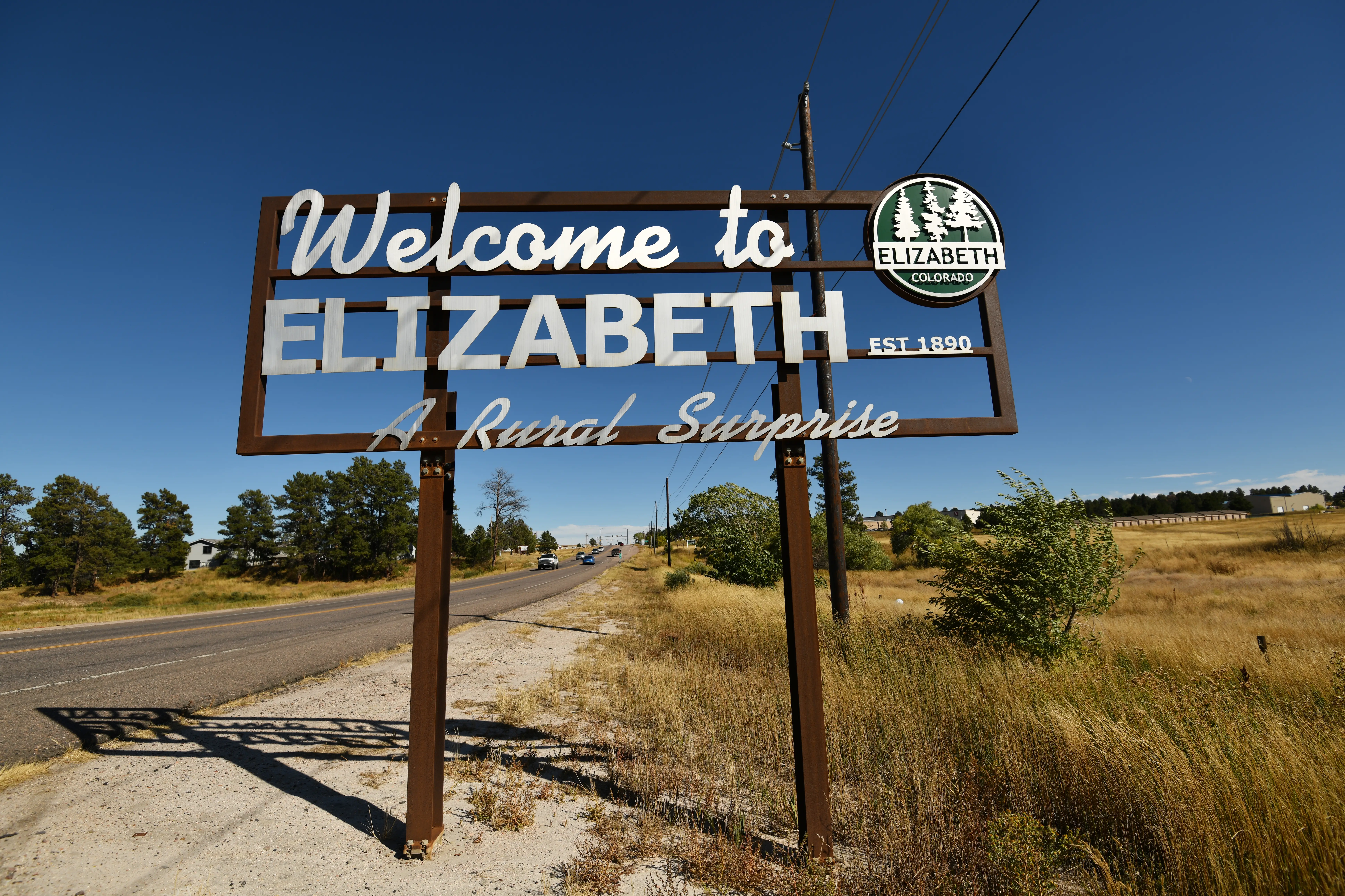 Roadside welcome sign for Elizabeth, Colorado, stands beside a rural highway under a clear blue sky.