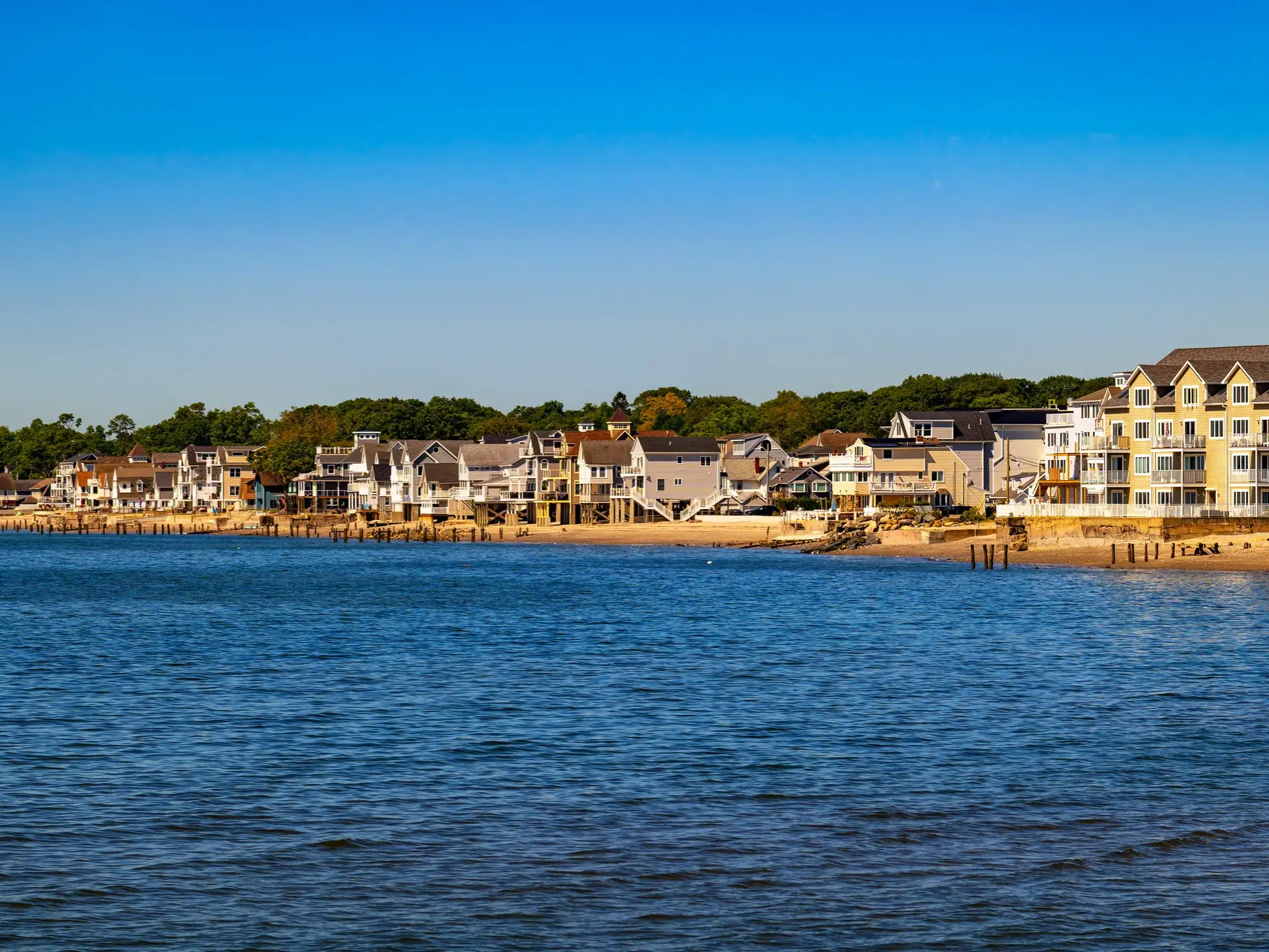 Waterfront houses line a sandy shoreline beside calm blue water under a clear sky.