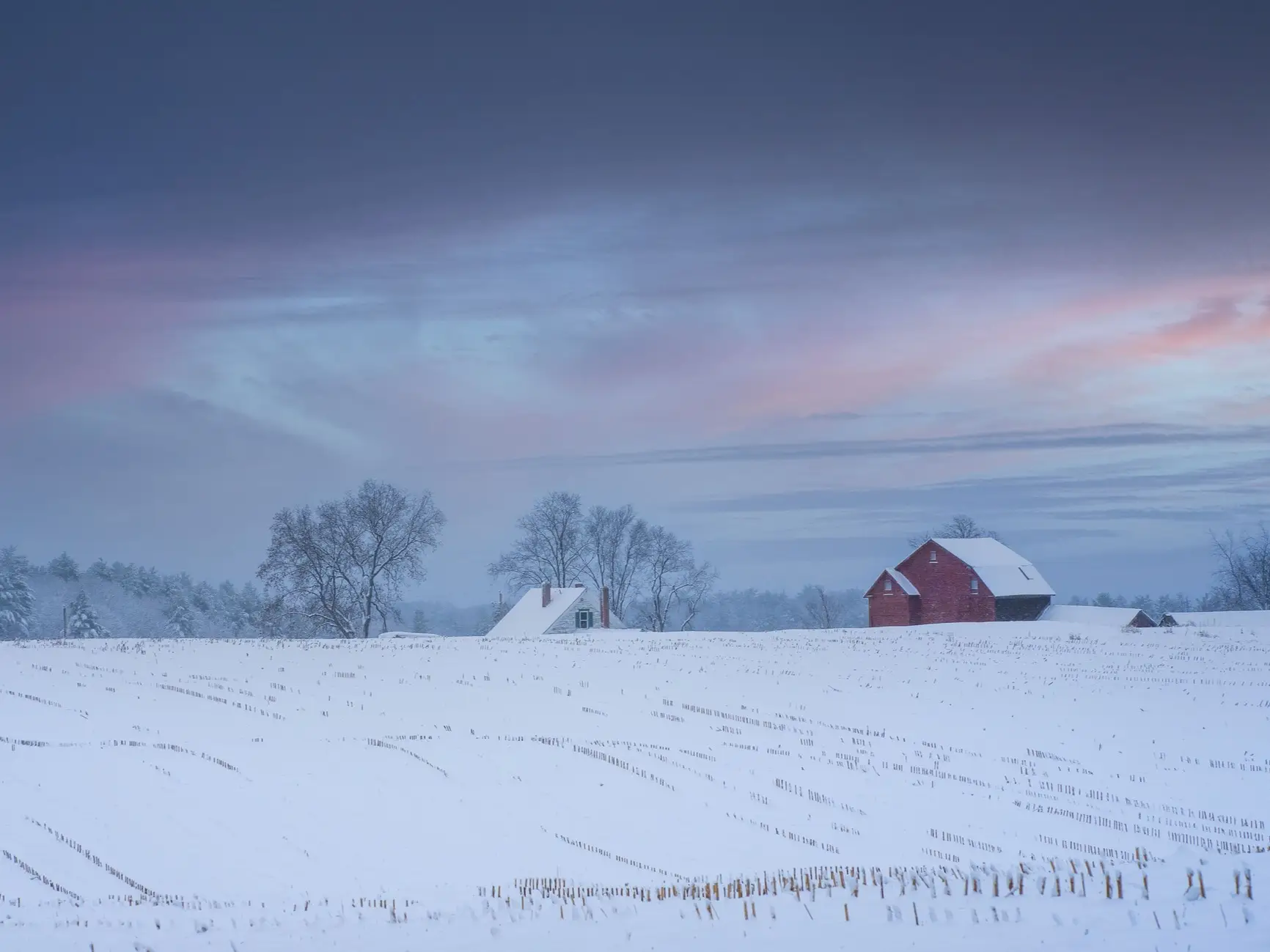 Snow-covered farm field with a red barn, small house, bare trees, and a pastel winter sky.