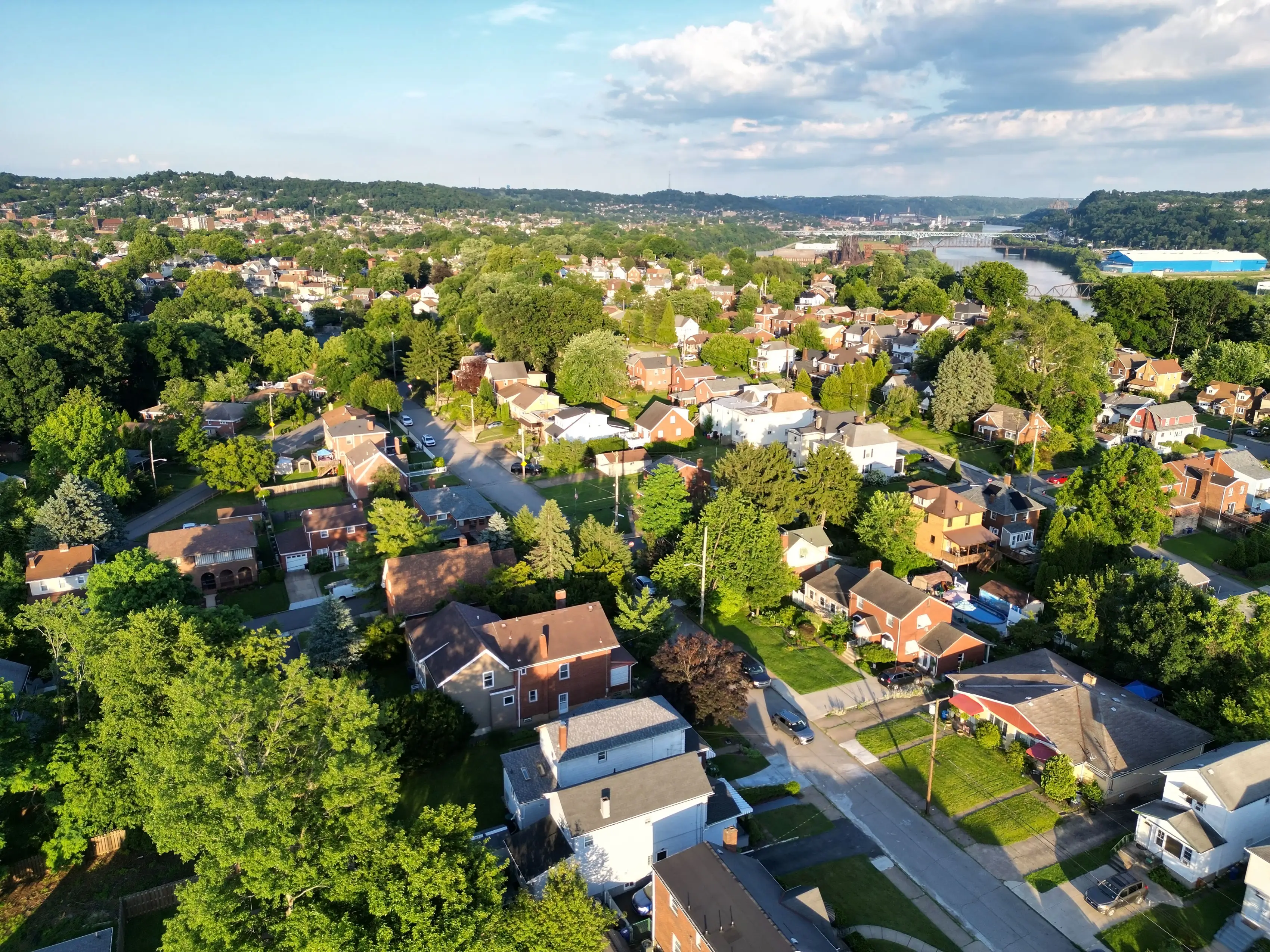 Aerial view of a leafy residential neighborhood with houses, streets, hills, and a river under a partly cloudy sky.